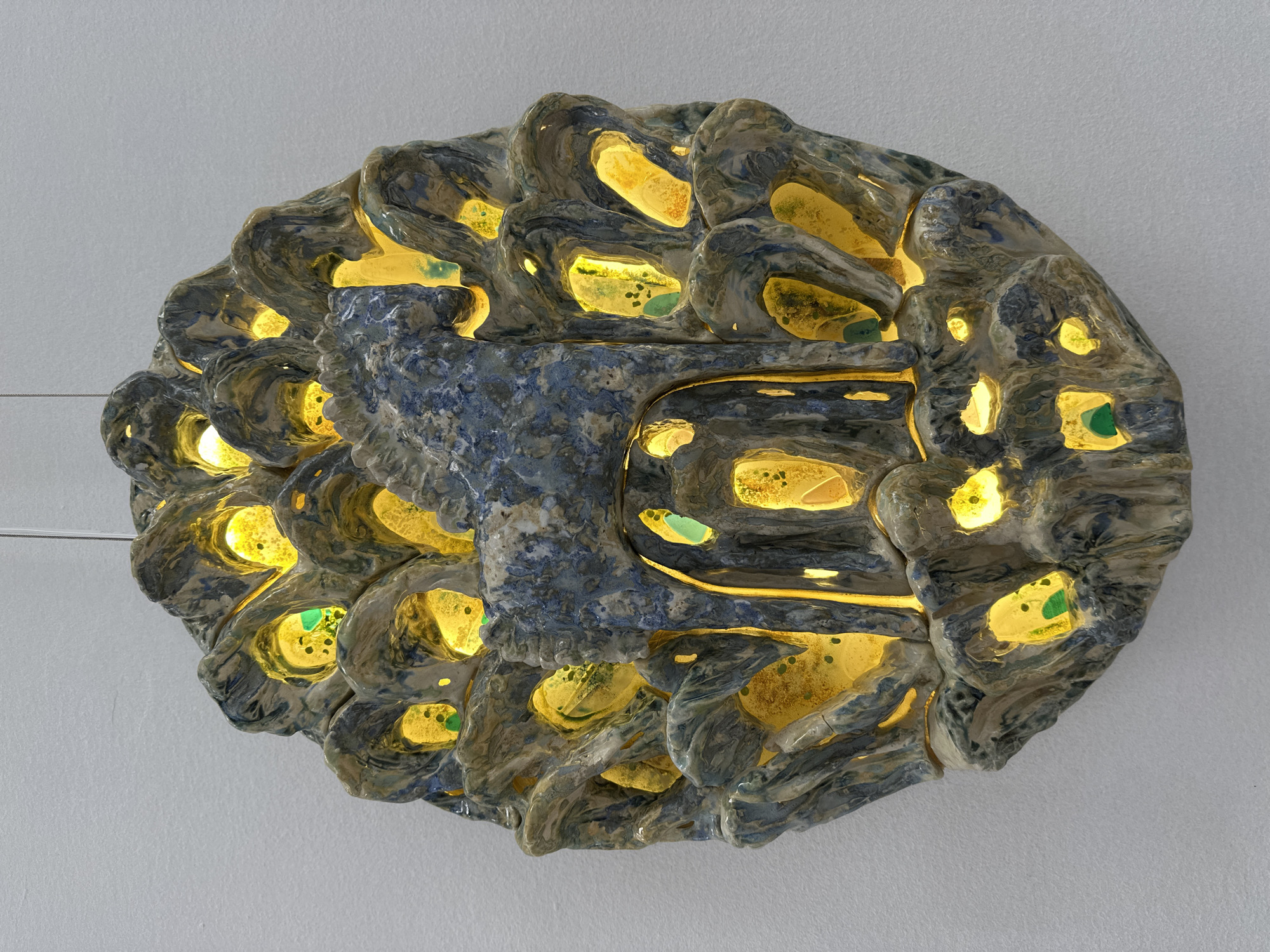 The media file presents an illuminated, abstract sculptural object, likely an art piece or decorative light fixture, set against a plain, light gray wall. The object is roughly oval-shaped with an irregular, organic, and highly textured surface, reminiscent of coral, a brain, or a complex natural formation.
It appears to be crafted from ceramic or a similar material, featuring a mottled coloration of blues, grays, creams, and subtle darker tones, with a slightly glossy or glazed finish. The surface is uneven, characterized by waves, ridges, and numerous deep cavities.
A warm, bright yellow light emanates from within these cavities, suggesting internal illumination. This light casts a glow on the surrounding textured surfaces and creates a striking contrast with the darker parts of the object. Within several of these glowing cavities, small, irregular pieces of translucent green material, possibly glass or polished stones, are visible. A more pronounced, somewhat smoother, blue-gray central section runs across the object, integrating with the more textured, undulating elements.
Thin wires, possibly for hanging or power, are visible on the left side of the object, extending from its edge. The plain background ensures that the intricate details and illumination of the object remain the sole focus. No activity is depicted, and there is no visible text.
