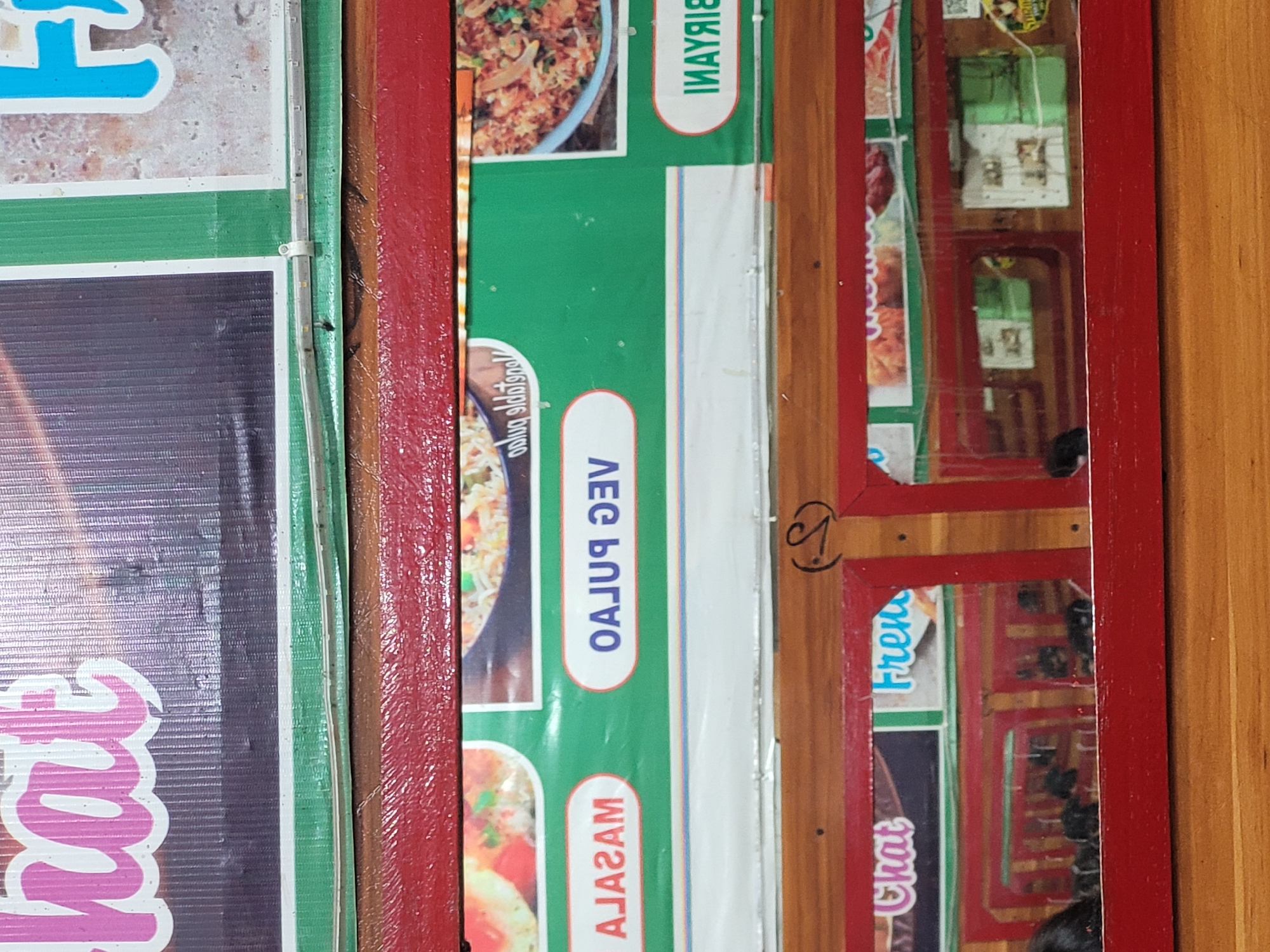 The image shows a close-up view of a food stall menu board and display case. The menu board features text and images of food items. One section of the menu board displays the words "MASALA" in white text within a red oval shape, above a picture of what appears to be a spicy dish. Another section shows "VEG PULAO" in white text within a red oval shape, with a picture of rice. The term "BIRYANI" is visible in white text within a red oval shape. In the lower part of the image, a red framed display case reveals reflections of the stall interior and some dark objects, possibly food items or equipment. The overall setting suggests a food vendor in Vashisht, India.