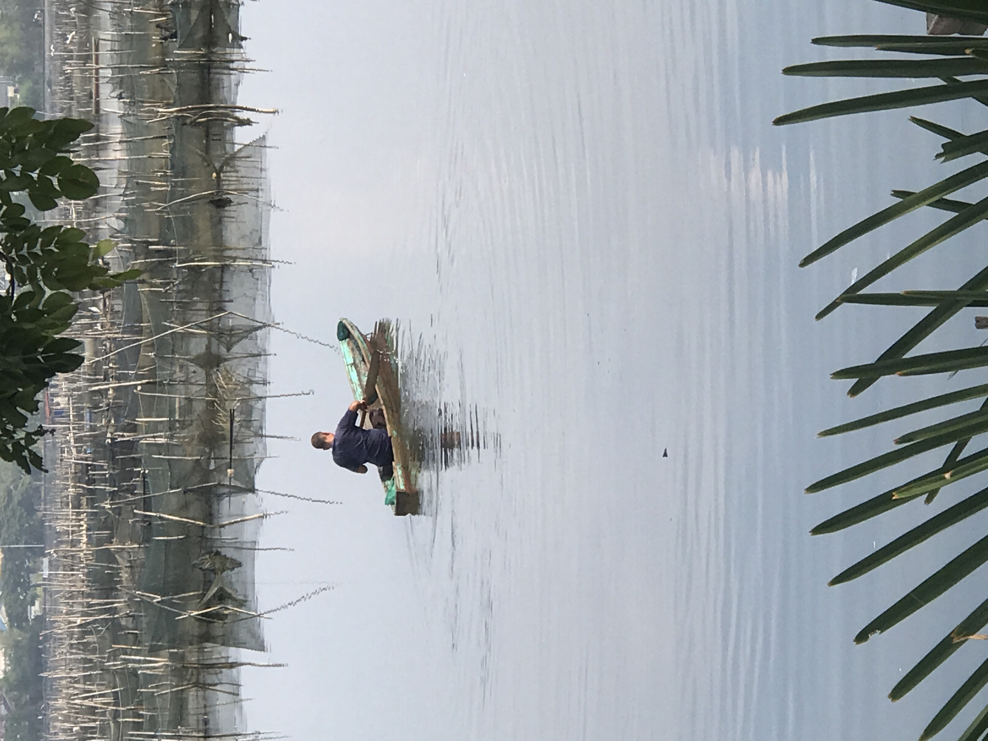 A man is on a wooden platform in a body of water, possibly a lake or river, near Lhokseumawe, Indonesia. The water has visible ripples. To the left are multiple wooden structures with netting, seemingly for fishing. Palm fronds are on the right. A tree is in the upper left. The sky is overcast, creating a muted light. The man is facing away from the viewer.