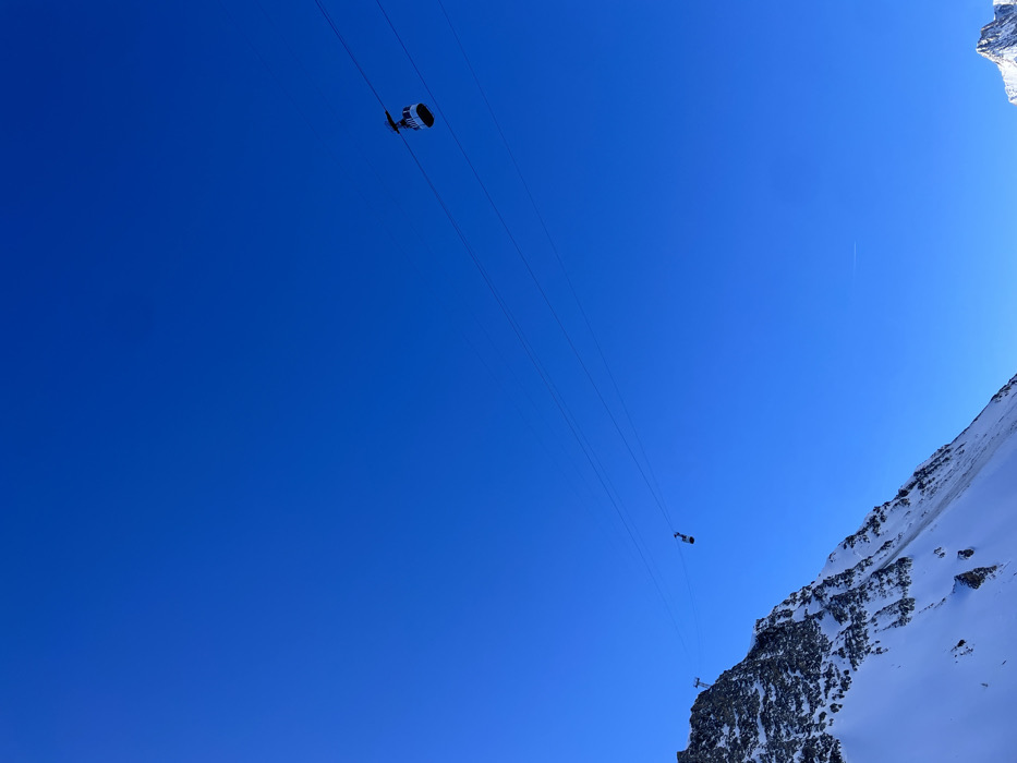 The image presents a striking view of a vibrant, clear blue sky dominating the upper and left portions of the frame, with a snow-covered mountain and rocky cliff face occupying the right side. Several parallel cable car lines stretch diagonally across the bright sky.
Two cable car cabins are visible on these lines. A larger, dark-colored cabin with prominent white components is positioned in the upper left quadrant, appearing to move along the cables. A second, smaller cabin can be seen further along the lines towards the mid-right, closer to the mountain's edge. A faint, thin white contrail, likely from a distant aircraft, is discernible in the upper right part of the sky.
The scene is set in a high-altitude mountain environment, characteristic of Courmayeur, Italy. It depicts a bright, sunny day with no clouds, suggesting excellent weather conditions. The strong sunlight creates clear visibility and highlights the snow and rock textures on the mountain. The perspective is from a low angle, looking upwards, emphasizing the vastness of the sky and the scale of the cable car system against the mountain backdrop. No visible text is present in the image.