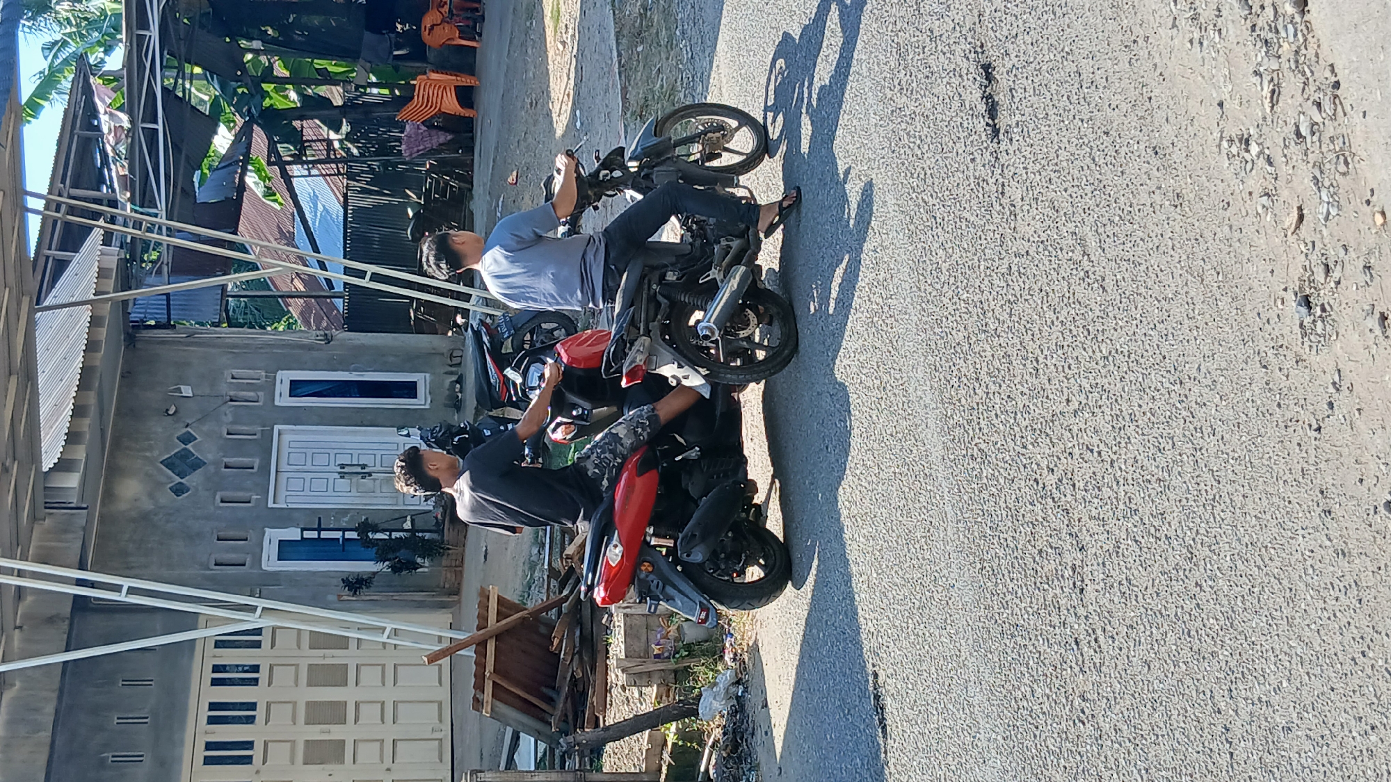 In Keudejungkagajah, Indonesia, two young men are seen from behind, each sitting on a motorcycle on a paved road. The man on the left, wearing a black t-shirt and camouflage shorts, is on a red motorcycle. The man on the right, dressed in a gray t-shirt and dark pants, is on a black motorcycle. They appear to be facing forward, possibly about to start riding. Behind them, a grey building with white-framed doors and windows is visible, along with some wooden planks and other debris. The environment suggests a rural or suburban setting with tropical vegetation visible in the background under a clear sky.