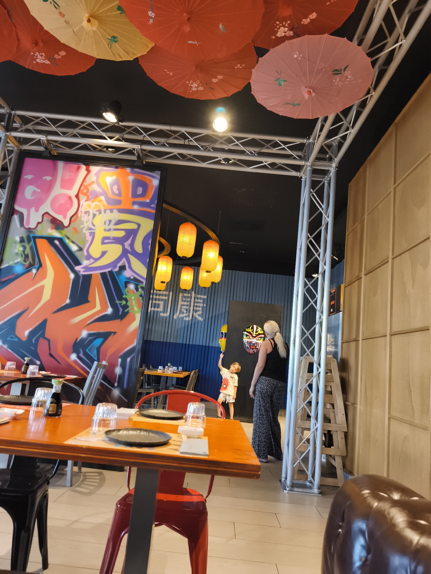 The image depicts the interior of a restaurant, likely a Japanese or Asian-themed establishment, with a focus on the dining area. Several tables are set with plates, glasses, and what appears to be soy sauce bottles. Red and black chairs surround some of the tables. Suspended above the dining area are several decorative paper umbrellas in shades of red and yellow, and some orange lanterns hang in the background. A large, colorful graffiti-style mural is visible on a wall to the left. In the mid-ground, a woman with blonde hair and a black top is standing next to a young child wearing a t-shirt. The child is reaching up towards a yellow object. A tall metal truss structure is present on the right side of the image, and a wall with a wooden paneling design is also visible in the background. The overall atmosphere suggests a lively and modern dining environment. The location in Livorno, Italy, is not explicitly indicated in the image itself, but could be inferred from external context.