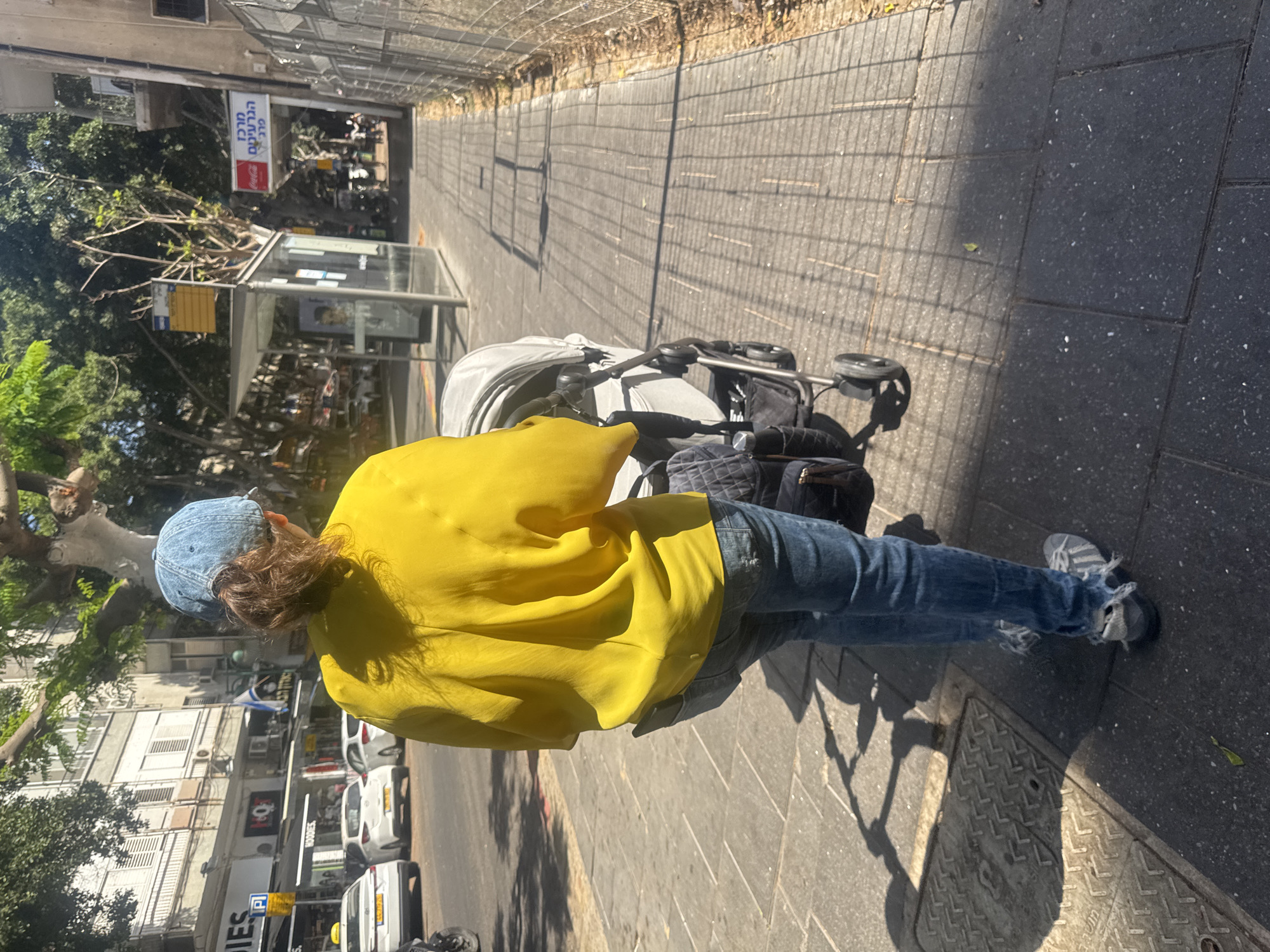 A person in a yellow shirt and blue jeans walks on a sidewalk in Ramat Gan, Israel, pushing a stroller. The sidewalk is made of gray concrete pavers and is bordered by a street with cars and buildings. A bus stop is visible in the background.