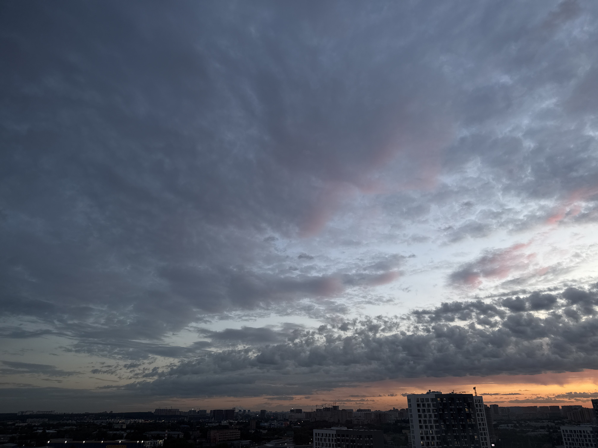 The image depicts a cityscape of Lyubertsy, Russia, during what appears to be sunset or twilight. The sky is filled with a dramatic display of clouds, ranging in color from dark grey and blue to lighter hues with hints of pink and orange near the horizon. These clouds are layered and textured, creating a sense of depth and movement.
Below the sky, the urban landscape of Lyubertsy is visible. Numerous buildings, including apartment complexes and industrial structures, form a dense silhouette against the sky. A prominent white building with dark-tinted windows is visible in the foreground to the right, featuring a construction crane on its roof. In the distance, the city stretches out, with lights beginning to illuminate some of the structures. The overall impression is one of an urban environment under a vast, atmospheric sky. No people or specific actions are discernible in the image.