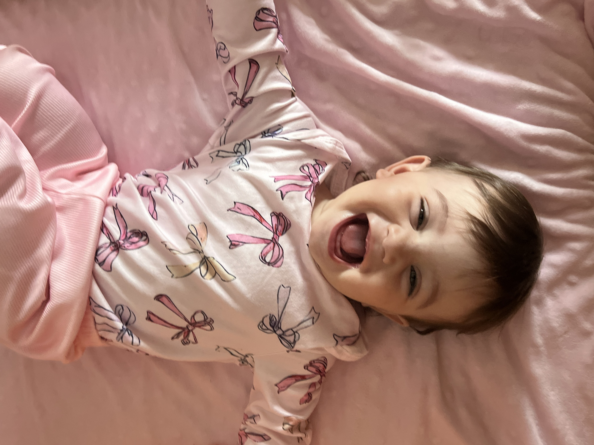A baby wearing pink clothing with a bow pattern lies on a pink blanket. The baby is smiling with its mouth open. The location is not discernible.