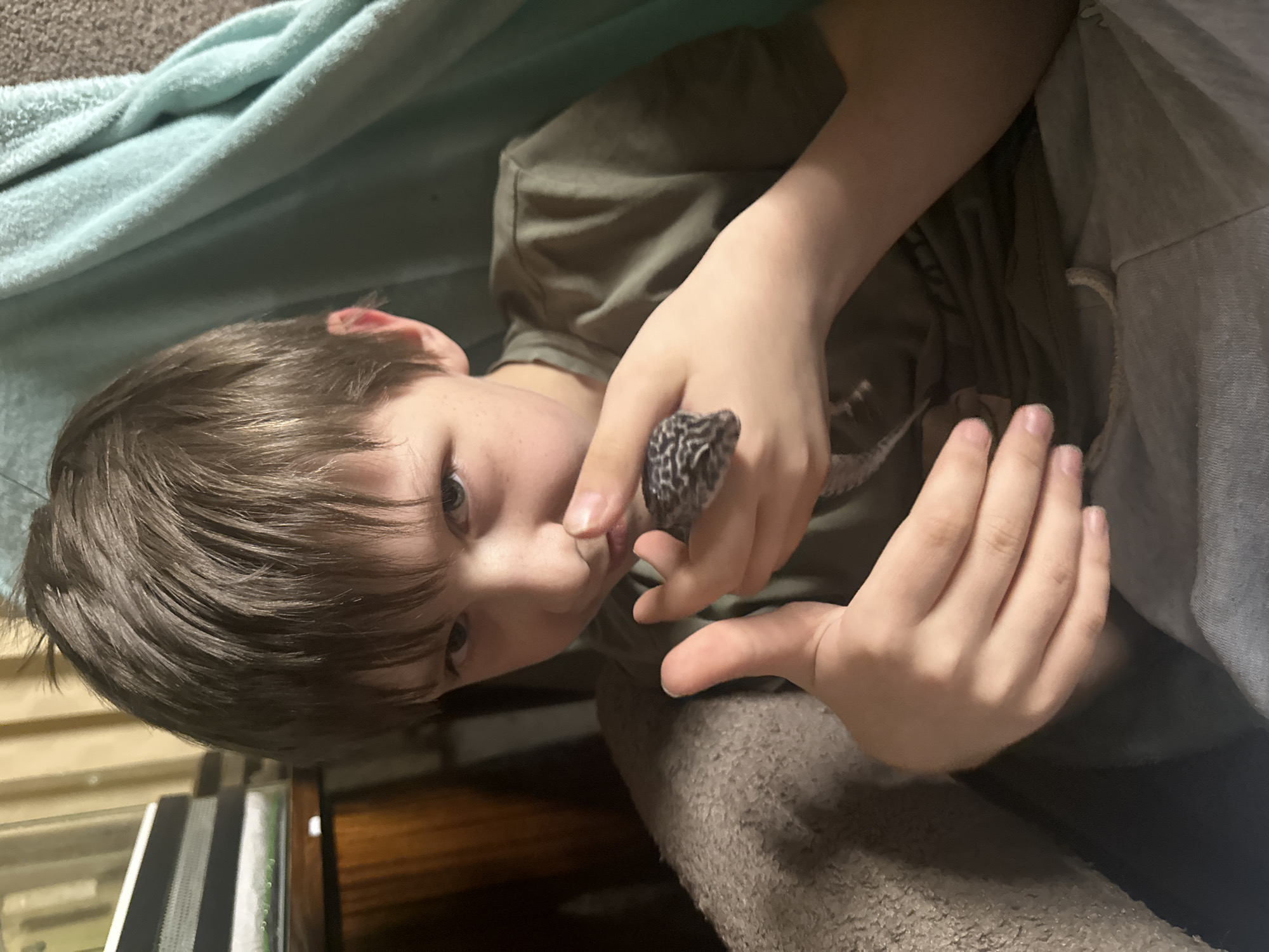 A young boy with brown hair is holding a leopard gecko in his hands. The boy is lying on a surface with a light blue blanket in the background and a brown surface below. The gecko is black and grey. The location is not discernible from the image.