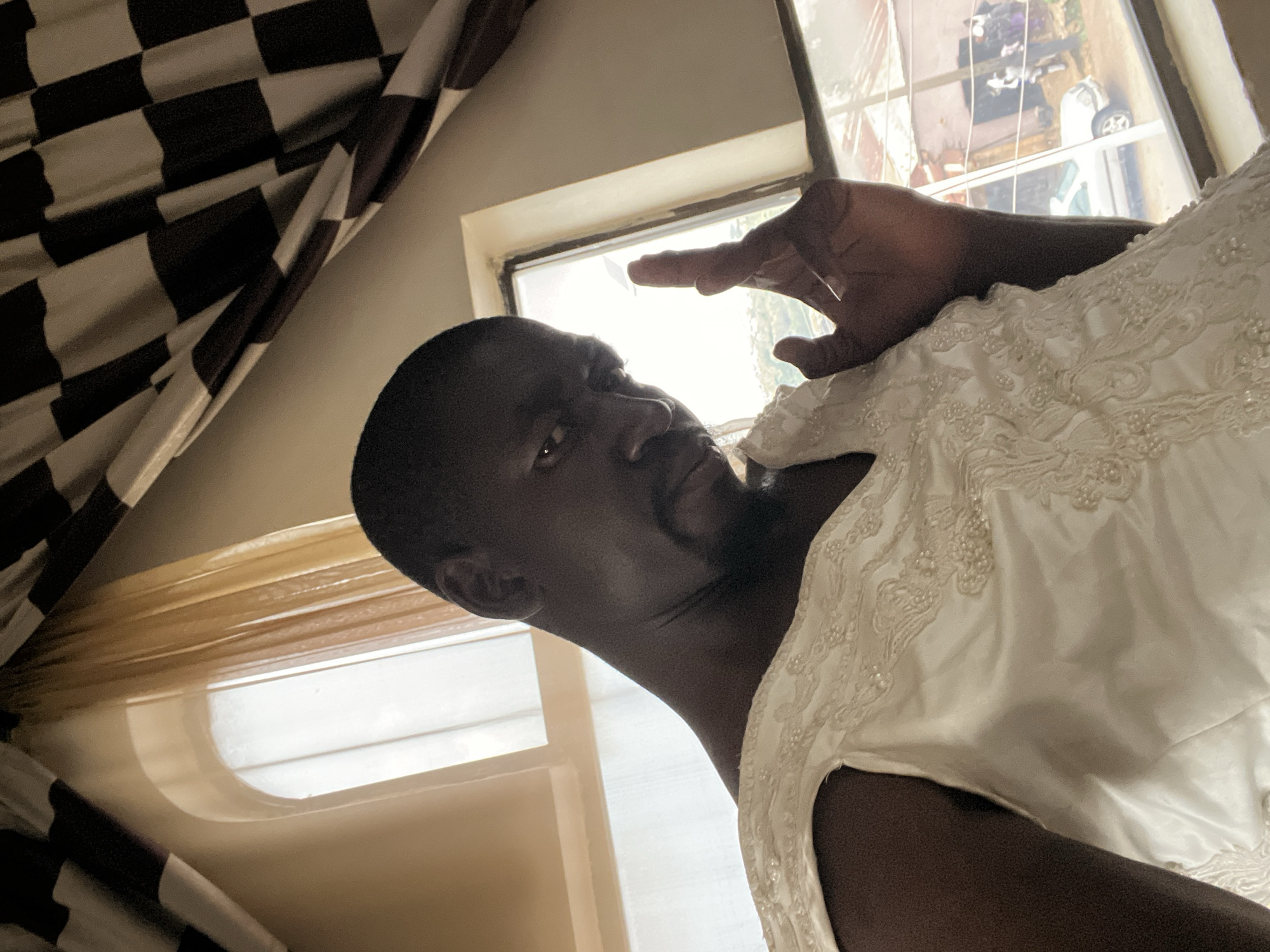 A medium shot captures a dark-skinned male indoors, wearing a white, beaded wedding dress. He is positioned in the foreground, angled slightly to his left, with his face turned towards the camera and his left hand raised with fingers extended. The man has short, dark hair and is looking directly at the viewer with a serious expression. The background reveals a window with light filtering through, and beyond the window, a blurred scene of buildings, a white car, and several people outdoors is visible, suggesting an urban environment. The window has a checkered curtain hanging above it. The overall lighting suggests it is daytime, with sunlight streaming through the window. The context of Jos, Nigeria is unknown and cannot be determined from the visual information.