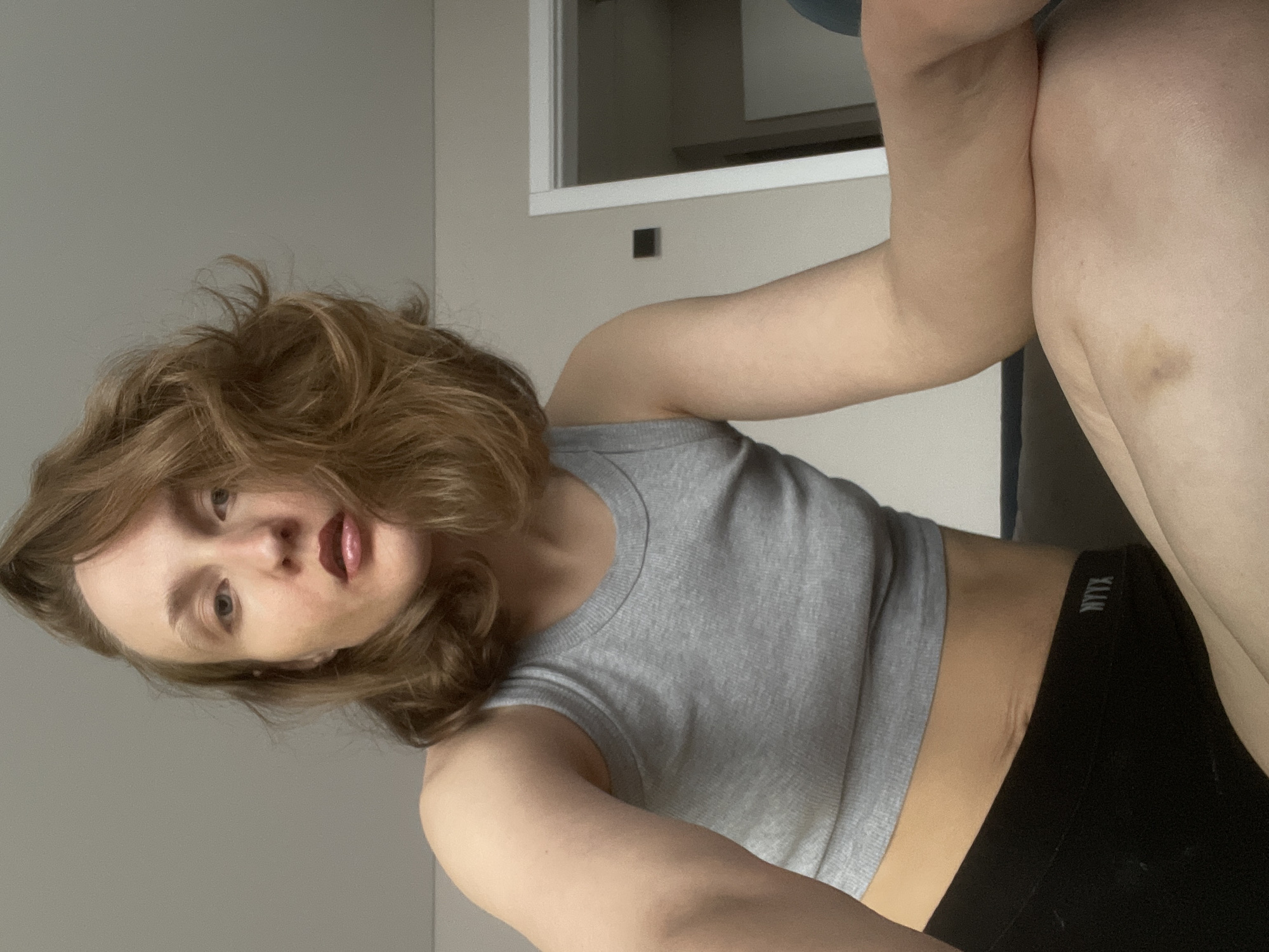 A woman is sitting indoors. She has shoulder-length wavy brown hair, light skin, and is wearing a gray tank top and black bottoms with the word "MYNX" printed on the waistband. There is a visible bruise on her thigh. The background includes a gray wall and a white cabinet. The image does not provide sufficient information to determine the location in Подшивалово, Russia.