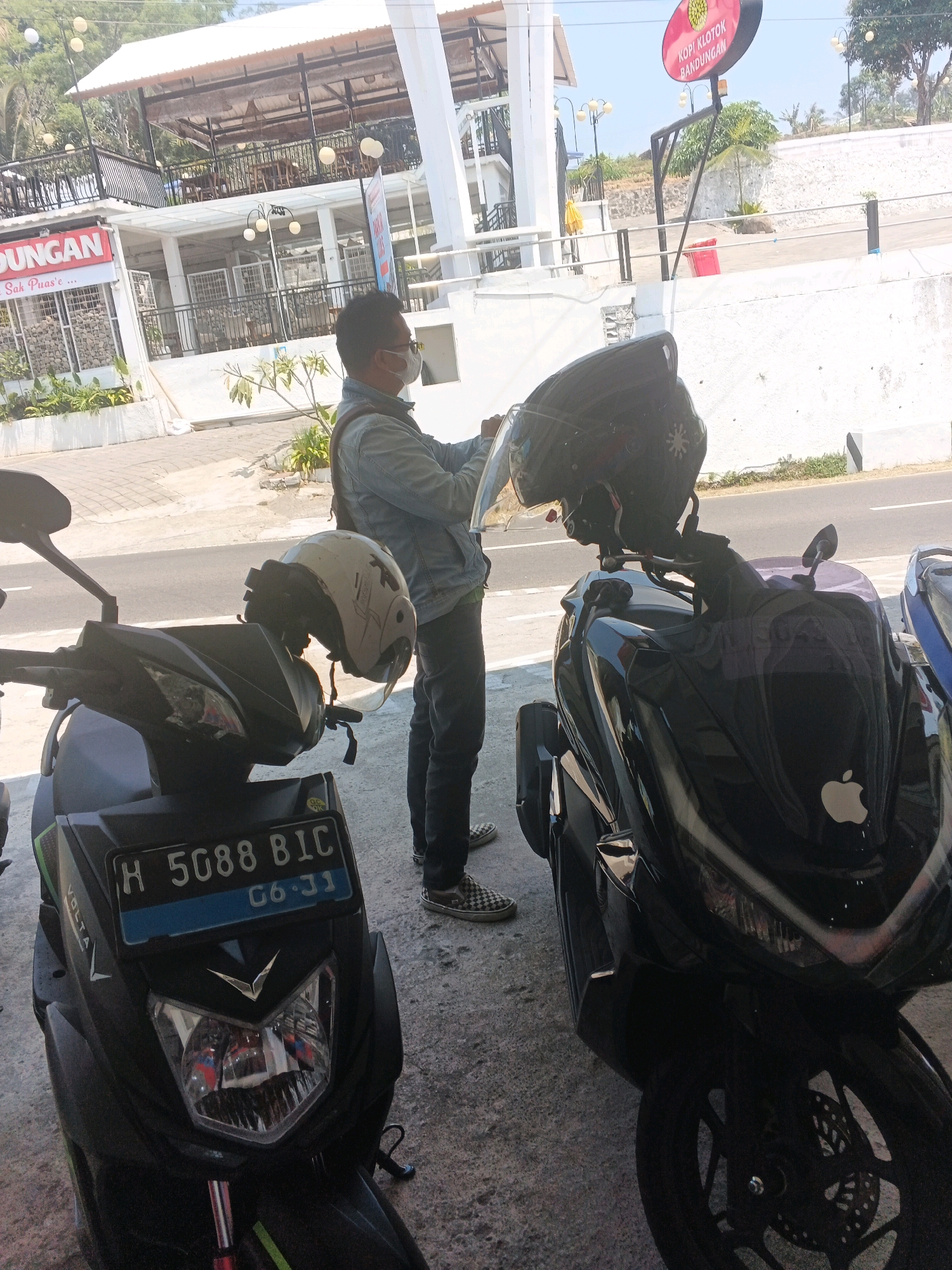 In Pakopen, Indonesia, a man wearing a blue denim jacket and a face mask stands between two parked motorcycles. He holds a clear helmet with a black helmet resting on top of it. The motorcycle in the foreground has a license plate that reads "H 5088 BIC" and "06-31". The background features a building with multiple levels and outdoor seating, suggesting a restaurant or cafe. A sign with red lettering that says "POPI KLOTOK BANDUNGAN" is visible. The scene is outdoors with a paved surface in the foreground and a road in the background.