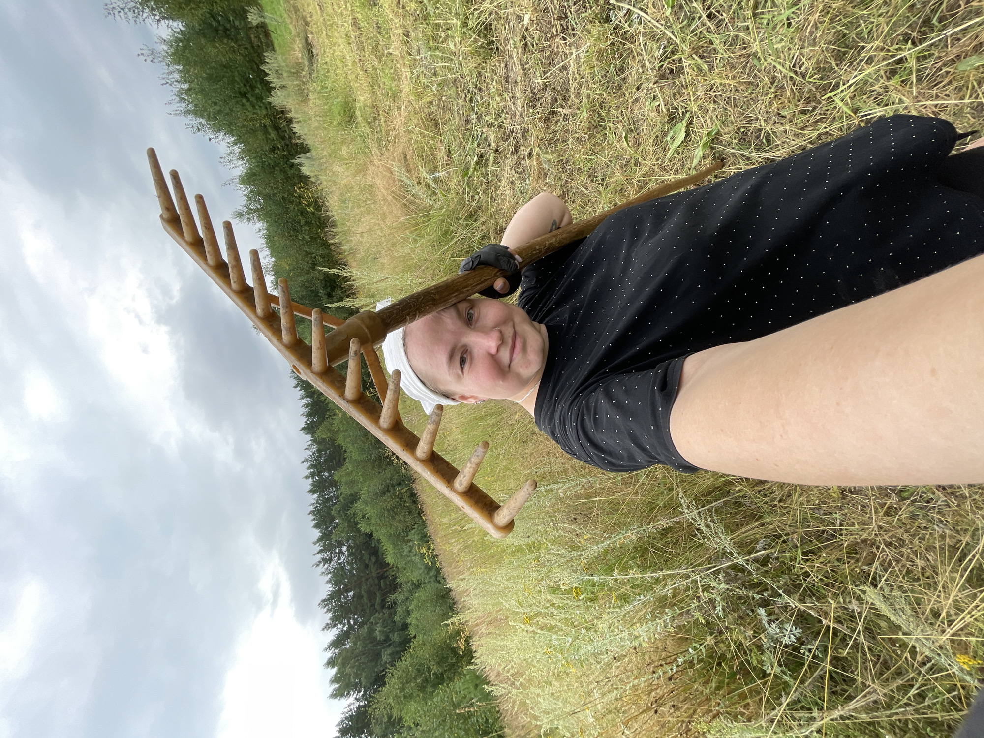 A woman, wearing a black t-shirt with white polka dots and a white headscarf, stands in a grassy field and holds a rake over her shoulder. She is looking at the camera and smiling. The field is dry and appears to have been recently mowed. In the background, there is a line of trees. The sky is cloudy. The location is likely a rural area outside of the city of Baldeyka, Russia, given the vegetation and open space.