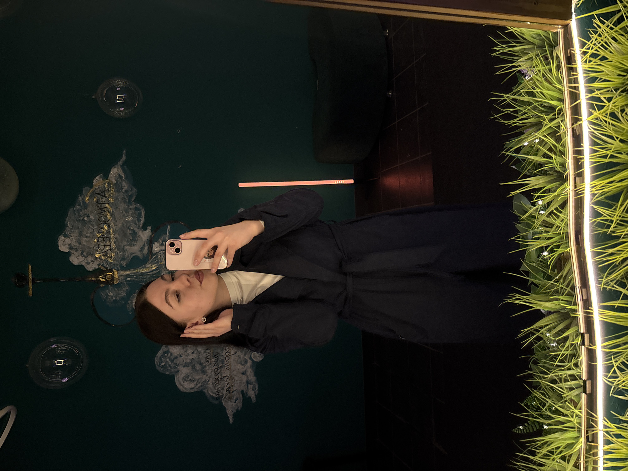 A young woman with long, dark hair is taking a selfie in front of a mirror. The mirror reflects a green wall with decorative elements, including stylized lamps, and what appears to be script. The setting is likely a decorative bathroom in Perm, Russia. The woman wears a dark wrap-style coat over a light-colored shirt. The right side of the mirror has a faux plant wall with integrated strip lighting. A dark interior background is also reflected in the mirror.