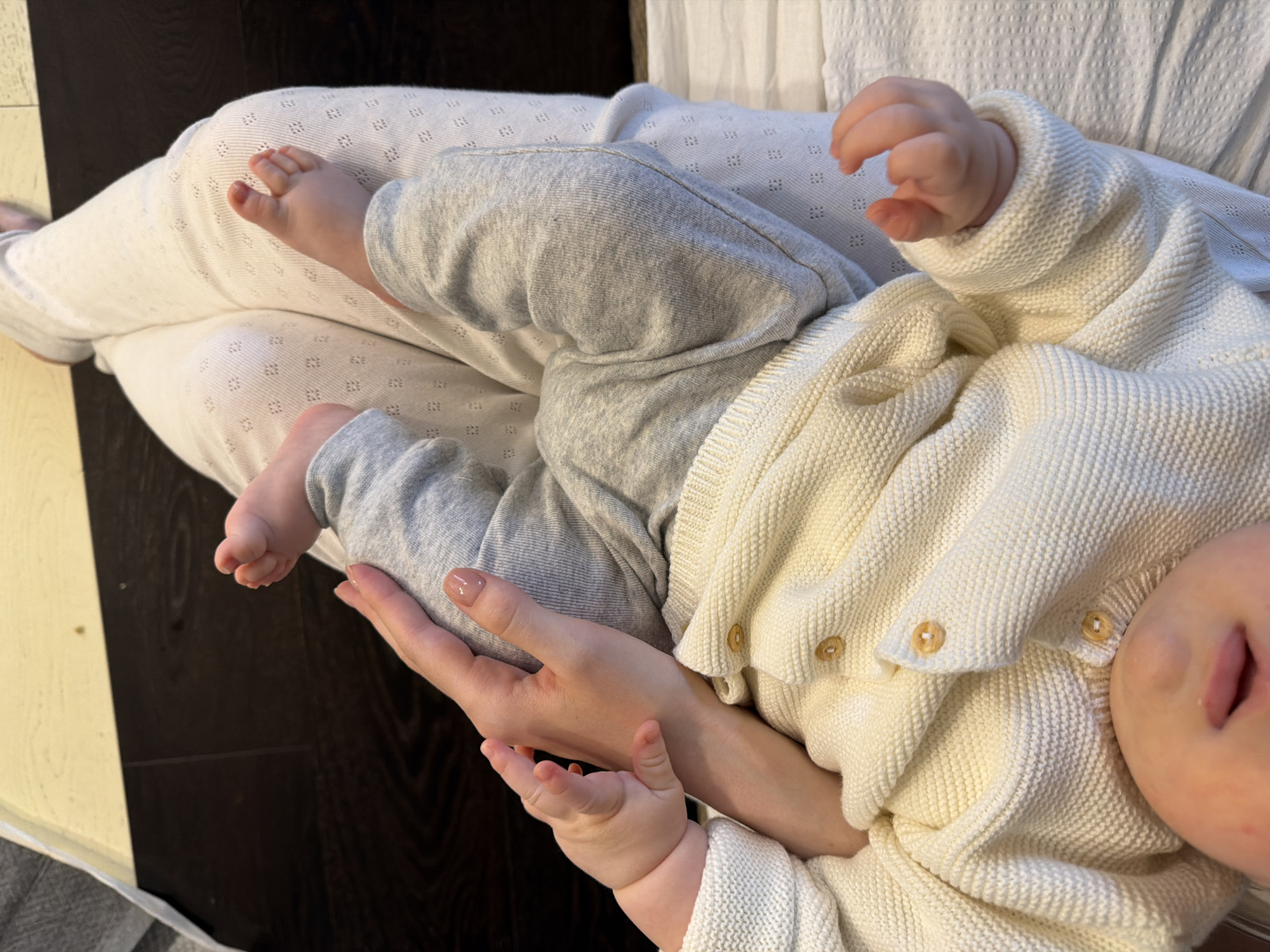 An overhead close-up shot shows an adult holding a baby, capturing a tender moment in an indoor setting in Khimki, Russia. The adult, whose legs are visible, is wearing white, textured leggings with a subtle square-dot pattern. One of the adult's hands is gently cradling the baby's back, revealing manicured fingernails with a light, natural polish.
The baby is dressed in light grey pants and a cream-colored, knitted cardigan featuring small, light brown buttons. The baby's plump hands and feet are visible, appearing soft and relaxed. One small foot rests on the adult's leg, while a tiny hand is clasped near the adult's hand. The baby's face is partially visible on the right side of the frame, with lips slightly parted, suggesting a state of relaxation or perhaps light sleep.
The background consists of dark wooden flooring or furniture, contrasted with lighter areas, possibly another piece of furniture or a wall, and a textured white fabric that might be a blanket or part of the bedding. The lighting appears soft and even, typical of an indoor environment, making it difficult to discern the specific time of day. No text is visible in the image. The overall impression is one of quiet affection and comfort between the adult and the baby.