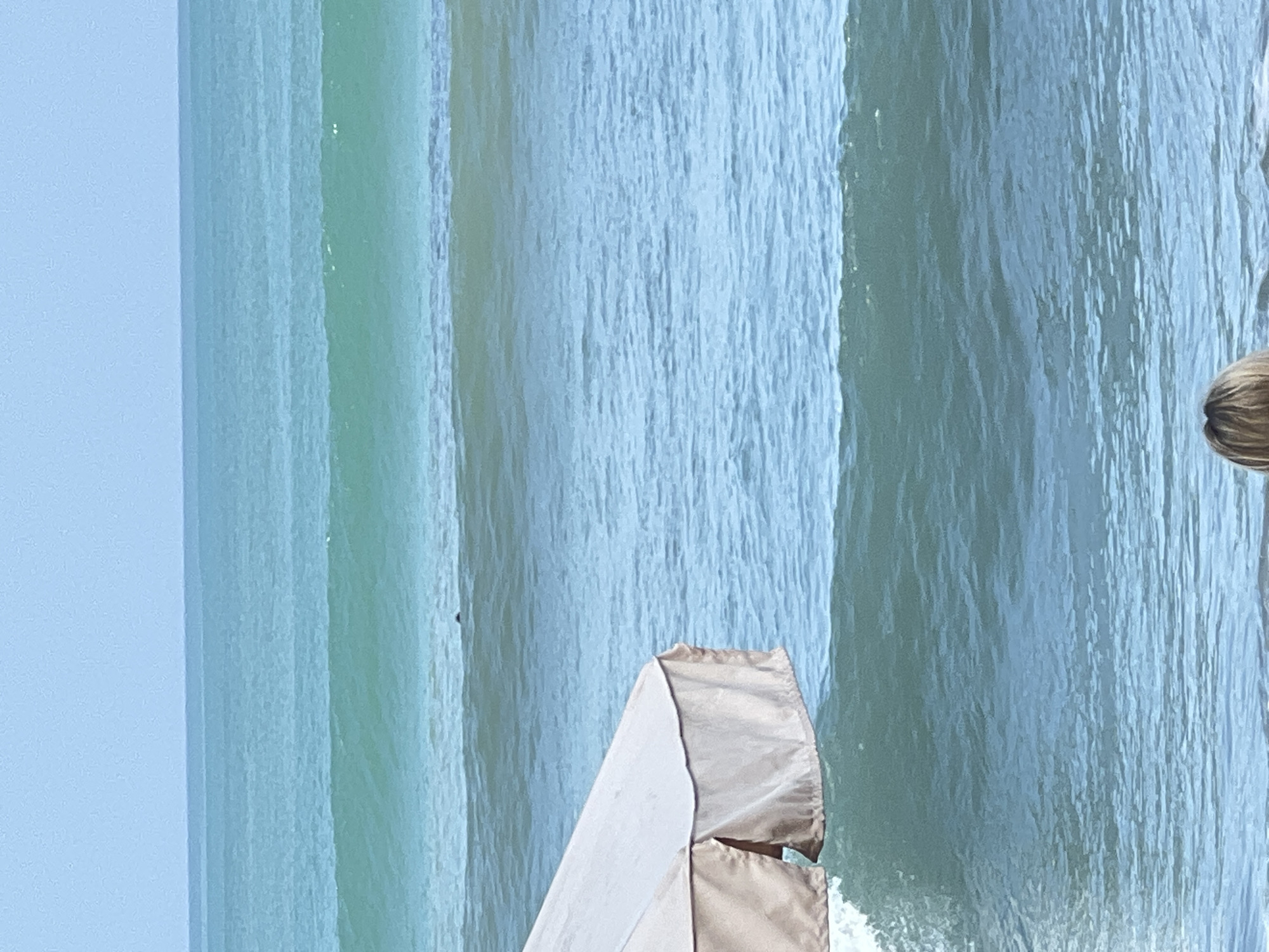 The image captures a serene ocean scene in Kuta, Indonesia. In the foreground, a portion of a light-colored beach umbrella is visible on the left side, casting a shadow on the water. The ocean stretches out, displaying multiple layers of turquoise waves. A solitary figure, possibly a surfer or swimmer, can be spotted in the distance amidst the waves, a small dark speck against the vast blue. The sky above is a pale, hazy blue, suggesting an overcast or early morning day. The overall impression is one of tranquility and the natural beauty of Kuta's coastline.