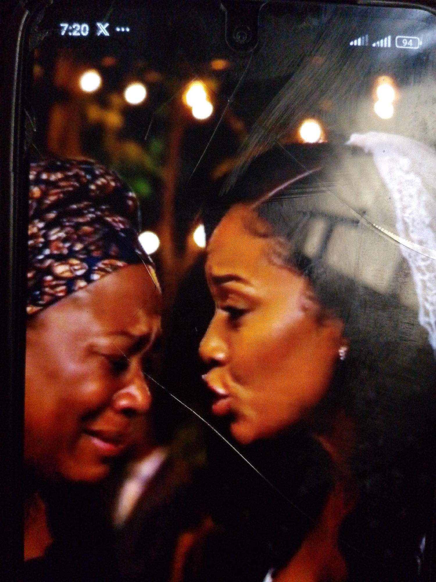 The image shows a close-up of two Black women's faces, captured on a phone screen which displays a cracked surface. The woman on the left wears a patterned headscarf and appears to be crying or expressing distress, with her eyes closed and mouth agape. The woman on the right has her eyes open and her mouth is slightly pursed, as if speaking or reacting. Her hair is styled in loose curls, and she wears a small earring. The background is dark and out of focus, with blurred lights that suggest an indoor setting, possibly a dimly lit event or a home environment. The phone screen itself displays the time "7:20" and signal strength indicators in the top left and right corners, along with a battery icon showing "94%". The overall scene conveys an emotional moment between the two individuals.