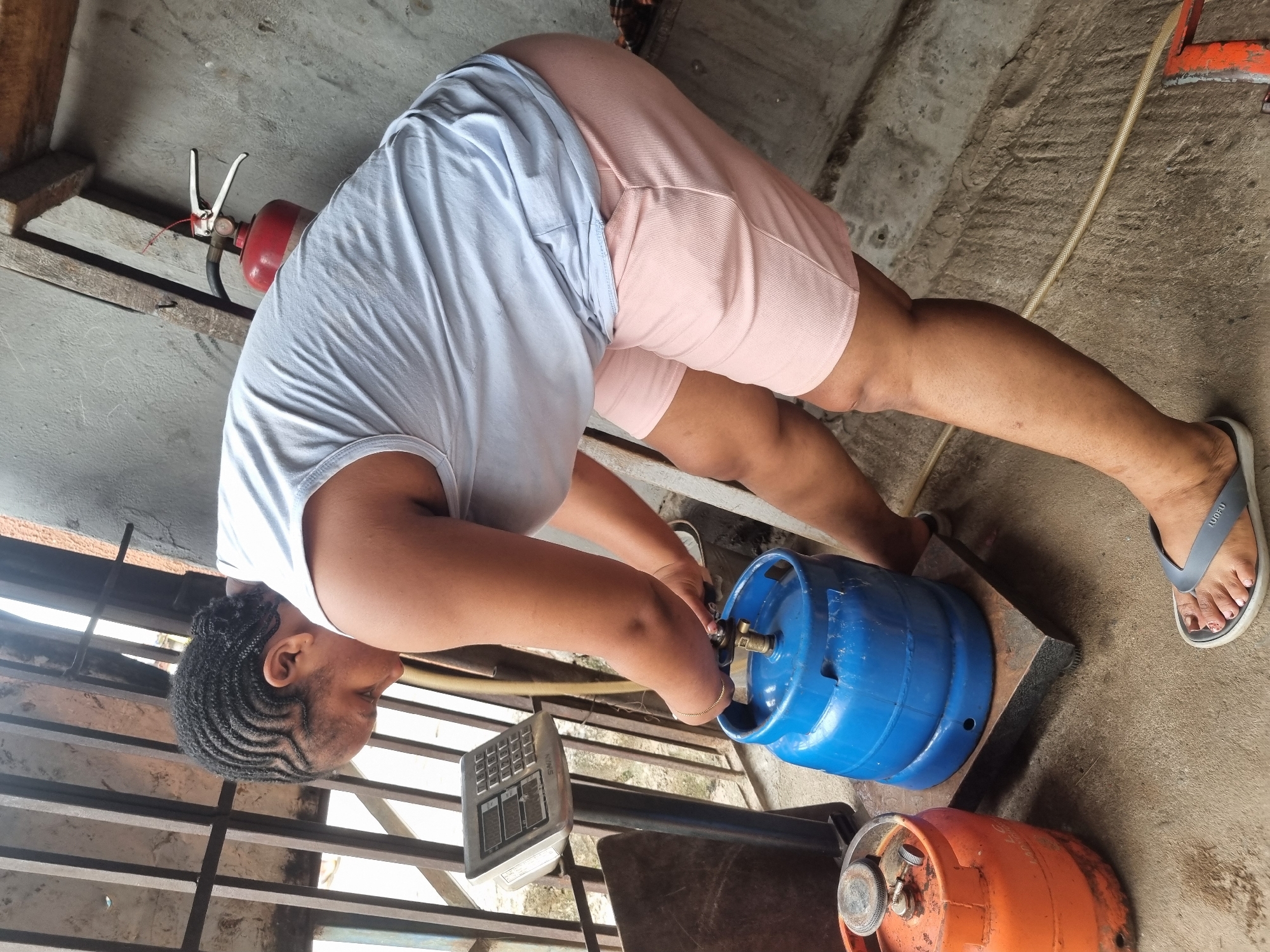 A woman is connecting a blue propane tank to a scale. She is wearing a light blue t-shirt and pink shorts, and is bent over, looking at the valve on the tank. The tank is on a metal base, and there is an orange propane tank on the ground nearby. A fire extinguisher is fixed to a wooden post above. The scene appears to be taking place outdoors, adjacent to a building, potentially within the city of Onitsha, Nigeria.