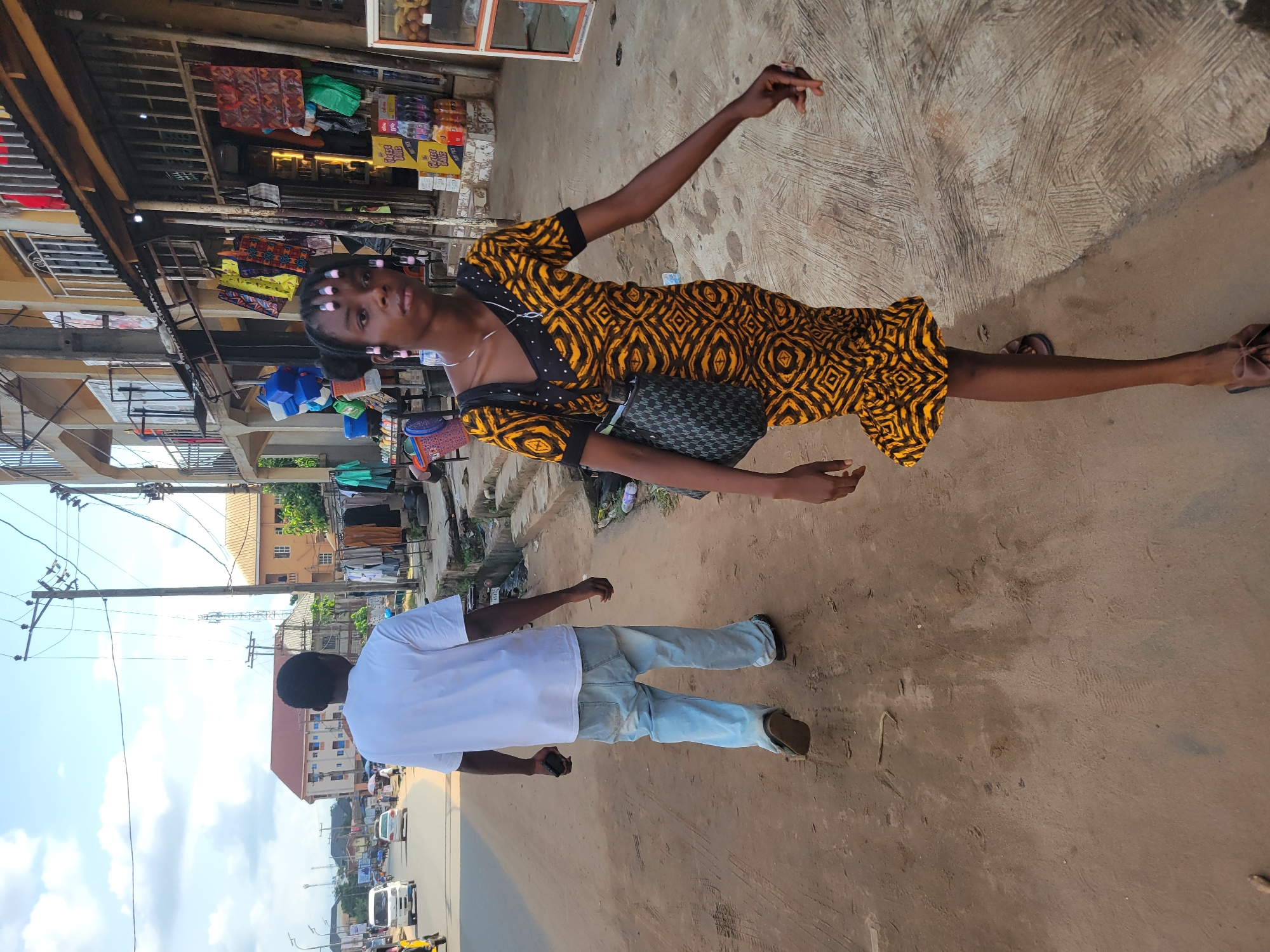 Two people are standing on a dirt path next to a road lined with shops. A young woman in a yellow and black patterned dress with pink rollers in her hair is posing with her arms outstretched. She is carrying a dark patterned bag on her shoulder. Behind her, a man in a white t-shirt and light blue jeans is facing away from the camera, holding a phone in his left hand. The scene appears to be a marketplace or a busy street in a town or city. There are shops with various goods displayed, including packaged drinks and bags. Overhead power lines are visible in the background, along with buildings and parked vehicles on the road. The lighting suggests it is daytime, with some shadows indicating sunlight. The overall atmosphere seems casual, possibly a moment captured during a walk or while shopping.