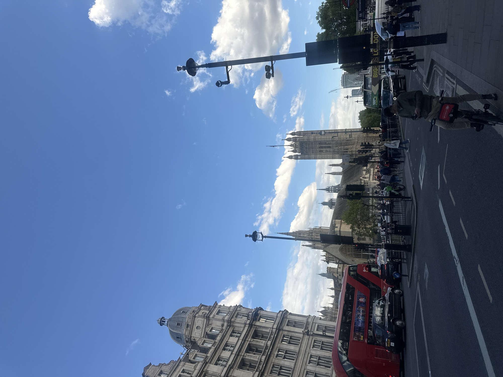 The image shows a street scene in London, United Kingdom, with the Houses of Parliament visible in the background. A red double-decker bus is present on the street, along with black taxis and other vehicles. Pedestrians are walking along the sidewalk. A cyclist is riding a bicycle on the right side of the image. The sky is blue with scattered clouds. A tall building with a dome is on the left side of the image. Streetlights with security cameras are also visible. The traffic is stationary.