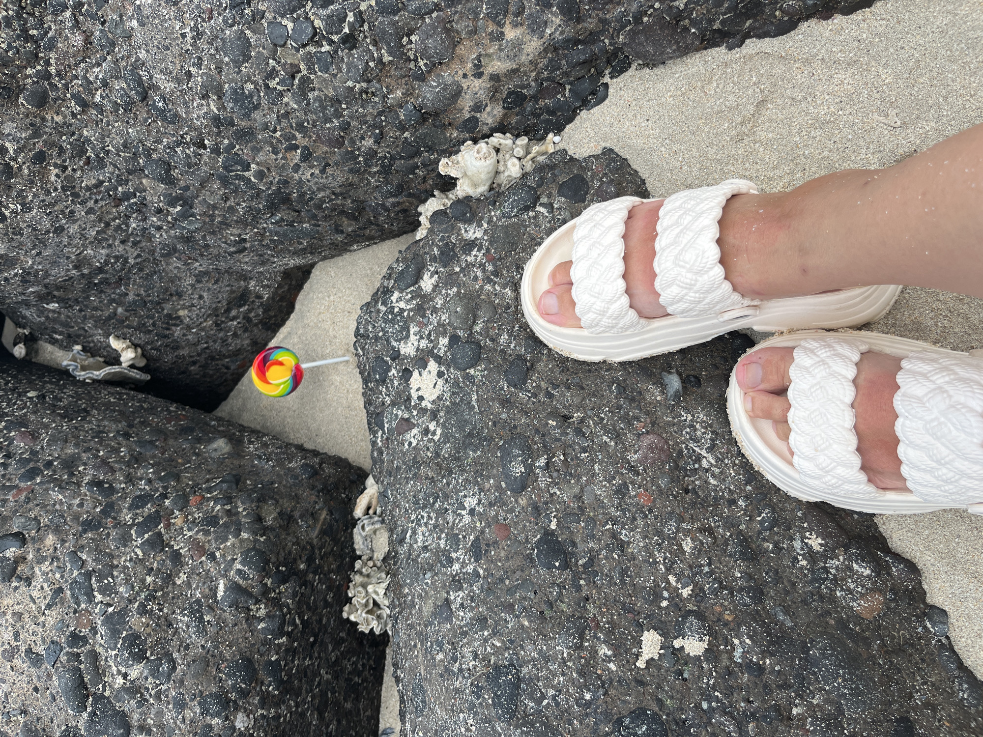 An overhead shot captures a pair of feet wearing white braided slides standing on a sandy beach, nestled between large, dark, rough-textured rocks. A colorful, round lollipop with red, yellow, green, and blue stripes is stuck upright in the sand between the rocks. Scattered white coral fragments are visible on and around the rocks, as well as small white specks that appear to be shells or debris on the sand. The lighting suggests an overcast day.