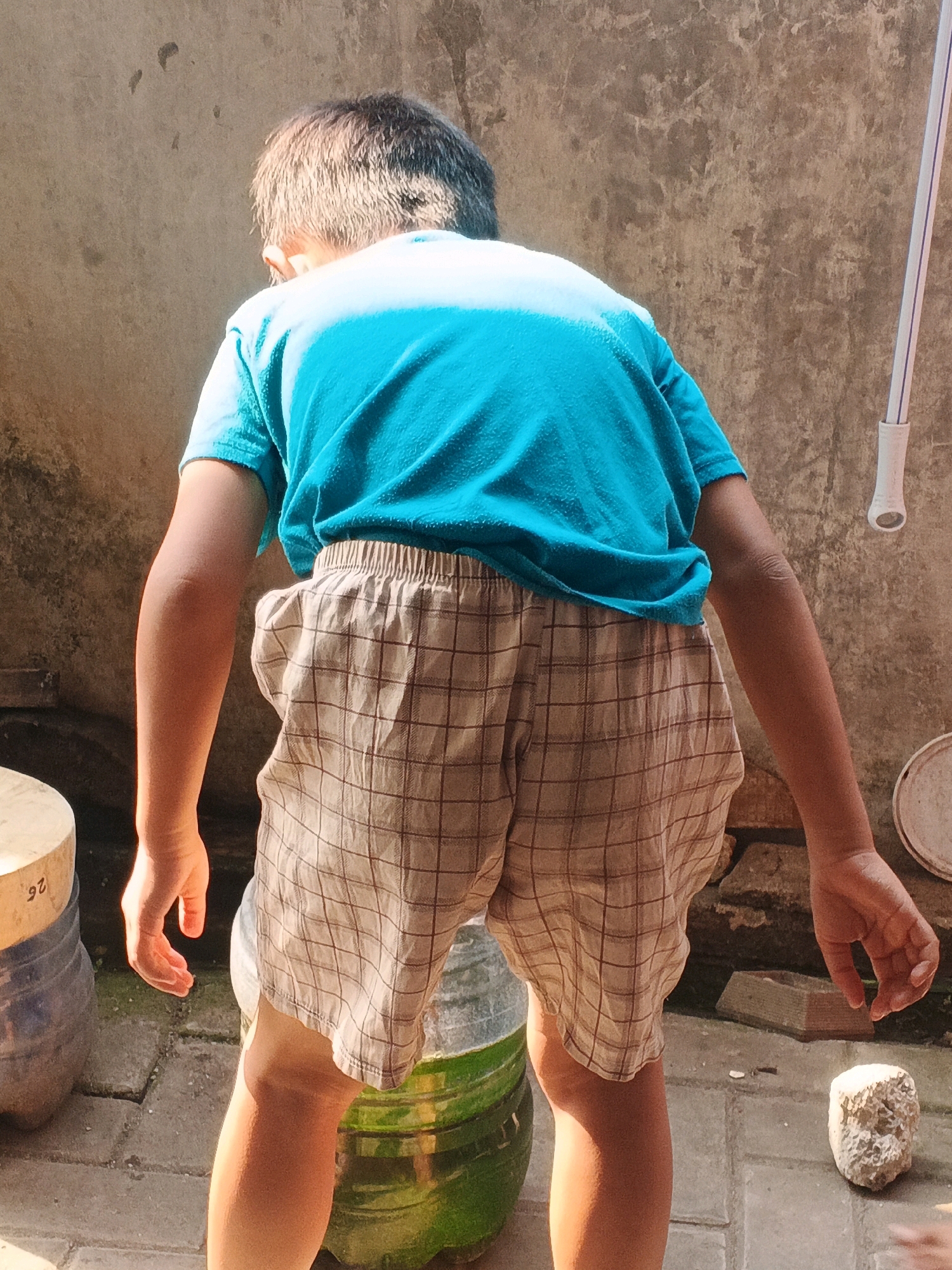 The image depicts a young boy, viewed from behind, wearing a bright blue t-shirt and plaid shorts. He is standing outdoors, possibly in a courtyard or alleyway, with a textured concrete wall as the backdrop. Sunlight casts shadows on the ground and the boy's body, indicating a bright, sunny day.
In the foreground, to the left, is a large cylindrical object, possibly a bucket or container, with a lighter colored top and a darker blue base. To the right of the boy, a portion of a green cylindrical object is visible, with horizontal bands. The ground is paved with square concrete tiles, some of which appear cracked. A small, rough, light-colored rock is also present on the ground to the right. In the background, the concrete wall shows signs of wear and possibly some plant growth. A white cylindrical object, possibly a pipe or part of a fixture, is attached to the wall on the right side of the image. The overall scene suggests a simple, outdoor urban or semi-urban environment.
Without additional context or specific landmarks, it is not possible to definitively identify the location as Kadu, Indonesia, based solely on the visual information in the image.