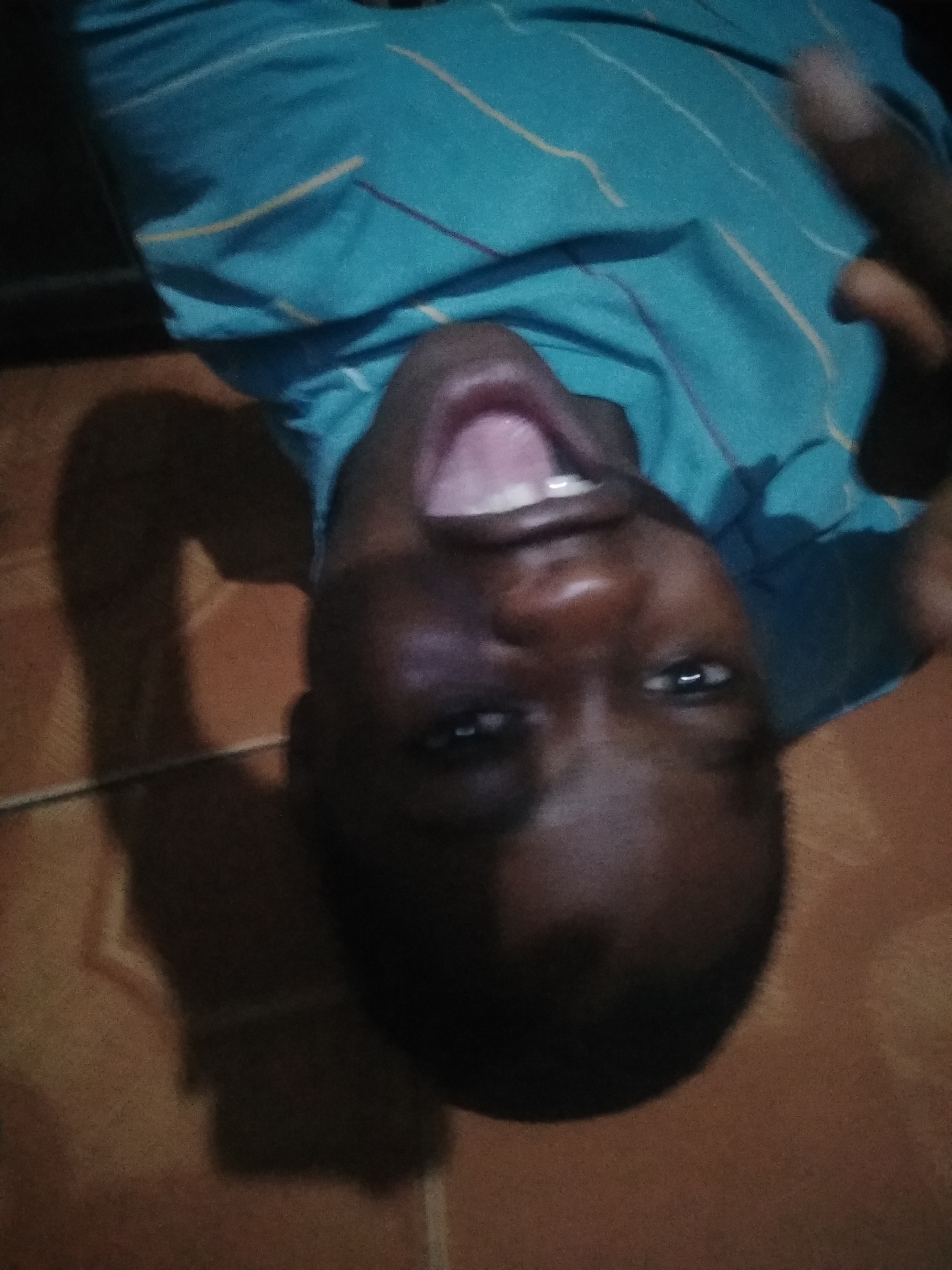A medium shot of a young, dark-skinned person lying on a tan tiled surface, possibly a floor. They are wearing a turquoise shirt with yellow and purple lines. Their mouth is open wide, and they are looking into the camera with their eyes slightly open. The image appears to have been rotated, as the person is upside down. There is a dark shadow cast on the tiled surface. The location and city are unknown.