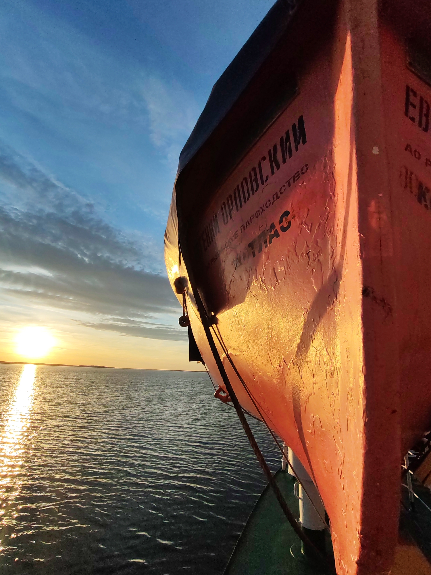 A close-up shot captures a lifeboat on a ship at sunset, against a backdrop of the ocean and sky. The lifeboat is painted in hues of red and orange, with Russian text visible on its side. The sunlight reflects brightly on the water, creating a shimmering effect. The sky is a blend of blue, orange, and gray, with scattered clouds. The photograph appears to be taken from the ship itself, offering a perspective that emphasizes the lifeboat and the natural setting.