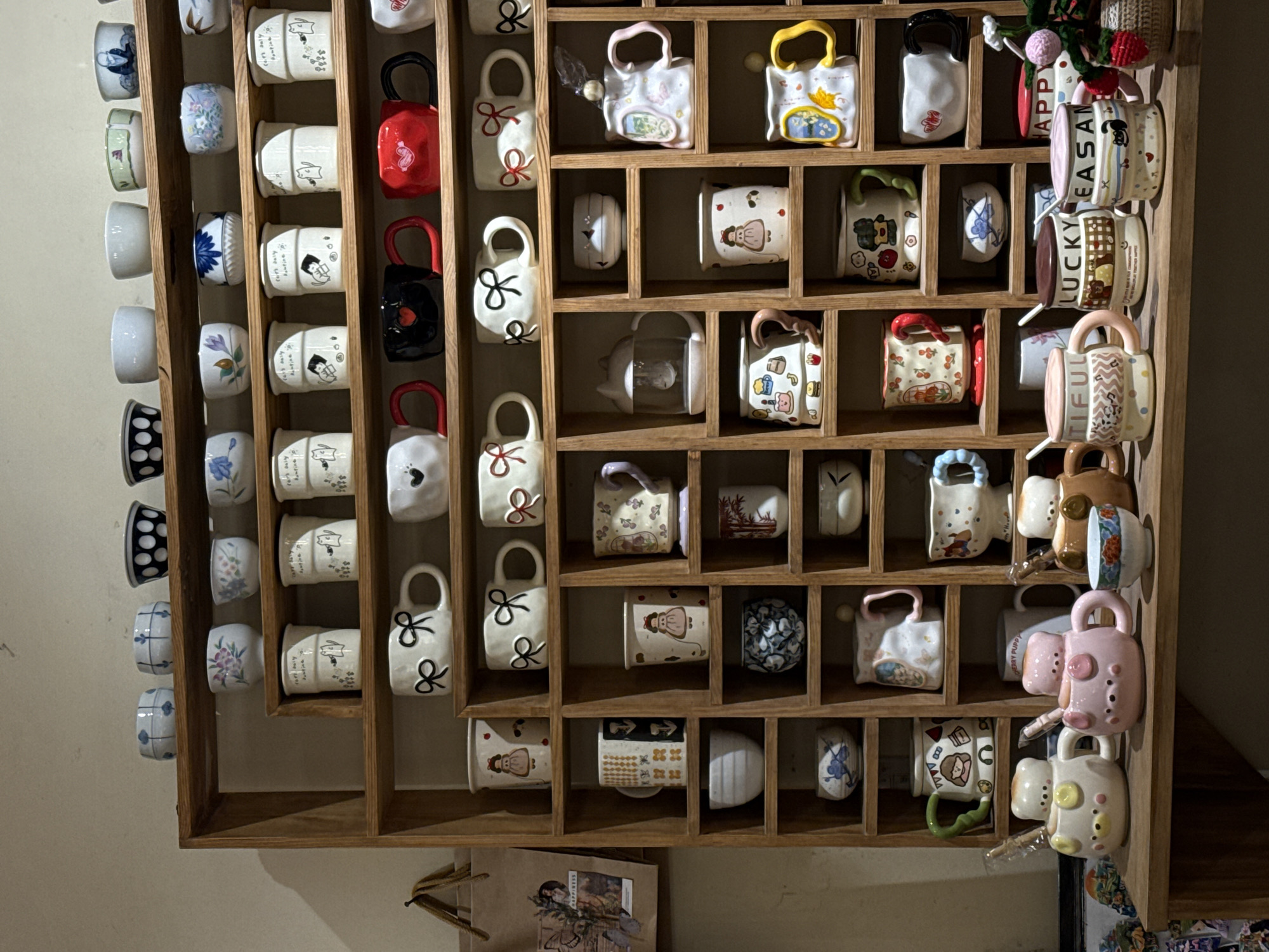 The image captures a wooden display shelf filled with a diverse collection of small ceramic items, primarily mugs and cups, likely presented for sale or as decorative pieces in Da Lat, Vietnam. The shelf is divided into numerous square and rectangular compartments, each housing one or more ceramic objects.
The ceramic items exhibit a wide variety of shapes, sizes, and designs. Many are small cylindrical mugs or cup-like vessels, while others are uniquely shaped like miniature handbags with prominent handles in colors such as red, black, pink, yellow, and white. The handles themselves vary, with some being simple loops, and others featuring decorative bows or patterns.
Designs on the ceramics include delicate floral patterns in blue, purple, and green hues; geometric shapes like polka dots and stripes; and whimsical illustrations of cartoon characters, animals, and stylized figures. Visible text on some mugs includes "2019," "HAPPY," "LUCKY," "EASAN," and "BEAUTIFUL," suggesting they might be souvenirs or celebratory items. One compartment on the top right holds a small vase-like object adorned with artificial red berries and knitted flowers.
Below the main display, a brown paper bag with indiscernible text and images is visible. The lighting is soft and ambient, suggesting an indoor setting, possibly a souvenir shop, cafe, or artisanal store common in a tourist destination like Da Lat. The overall impression is one of a charming and curated collection of handcrafted or mass-produced decorative ceramics.