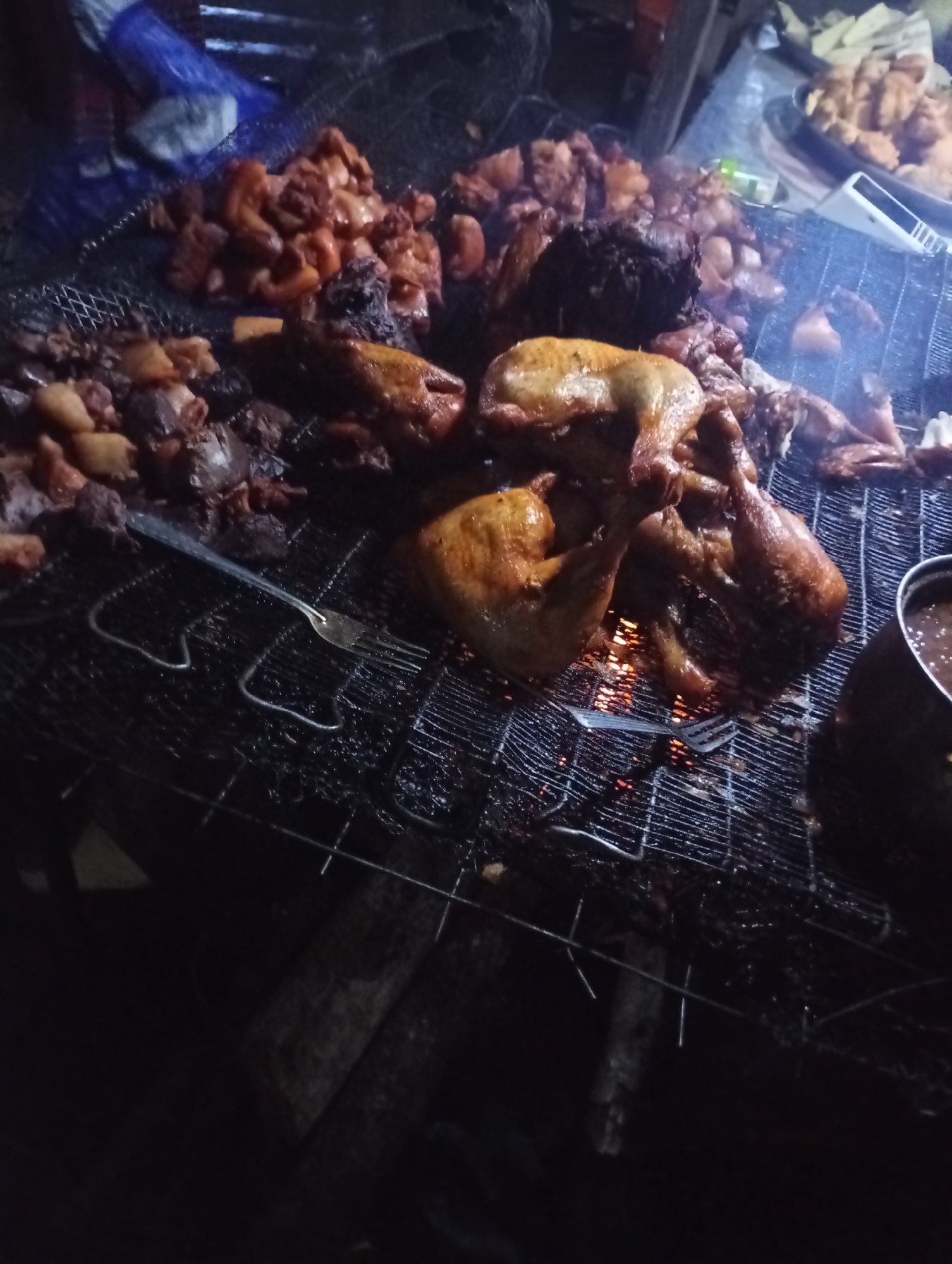 In an outdoor, low-light setting, a metal grill is filled with various pieces of meat being cooked over an open flame. The grill contains several chicken leg quarters, some appear to be fully cooked and browning, while others are still cooking. There are also piles of smaller, diced pieces of meat, likely pork or beef, scattered across the grill, some appearing fatty and charred. In the background, a large metal bowl filled with a dark sauce or gravy is visible on the right side of the grill. A metal fork is also present on the grill. The overall scene suggests a street food vendor preparing grilled meats. The exact location within the city is unknown.