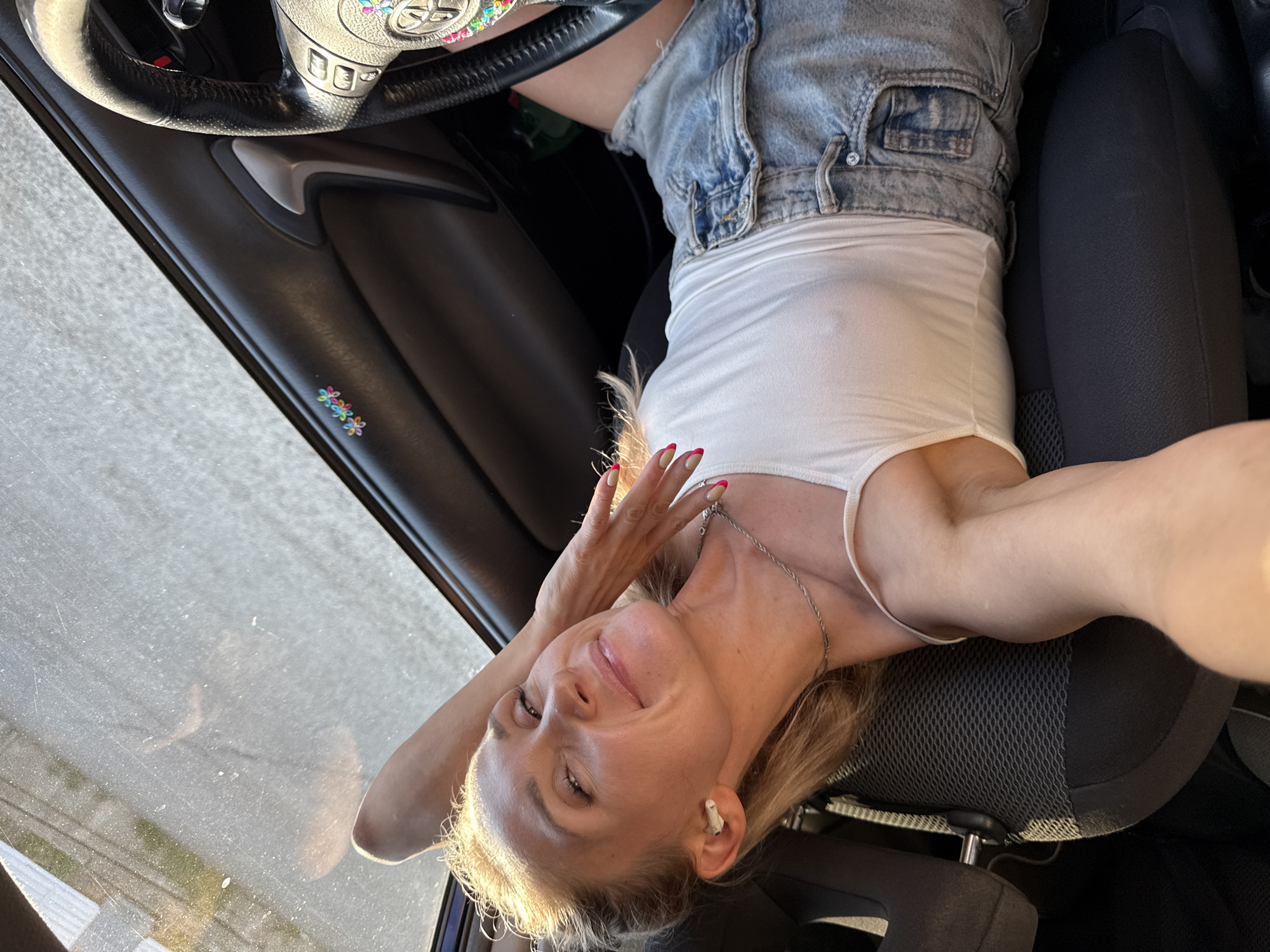 A woman is sitting in the driver's seat of a car. She is wearing a white tank top and denim shorts. Her hair is blonde, and she has a silver necklace. She is wearing an earbud in her right ear. Her hand is raised near her face, and she is smiling. The car's steering wheel is visible with decorative flower-like stickers. There is a sticker of small flowers on the car door. The car is parked on a paved surface, likely a street or parking lot. The location is Chaikovsky, Russia.