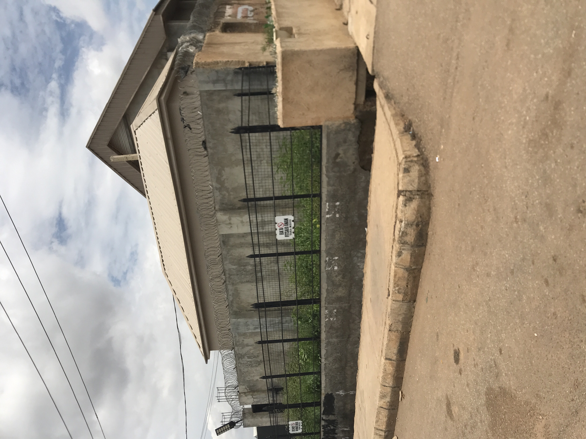 The image depicts an outdoor scene in Rafin Par, Nigeria, featuring a building and an adjacent street under an overcast sky with patches of blue. The primary subject is a two-story building with a light brown corrugated metal roof, exhibiting a prominent triangular gable on its upper section. The property is secured by a tall concrete perimeter wall, topped with multiple coils of razor wire. Below the razor wire, a black metal grid fence is installed above a concrete base, behind which green vegetation is visible. Two white rectangular signs are affixed to this fence. The left sign, partially obscured, reads "DO NOT DUMP REFUSE HERE" and "FINE N5000". The right sign clearly states "BAKI FITSARI ANAN" (Do not urinate here) and "TARA: N5000" (Fine: N5000), featuring a red prohibitory circle over a urinating figure. Overhead utility lines traverse the sky. A paved, light brown road, marked with stains, occupies the foreground, bordered by a concrete curb and an adjacent concrete drainage structure on the right. No individuals are present in the scene.