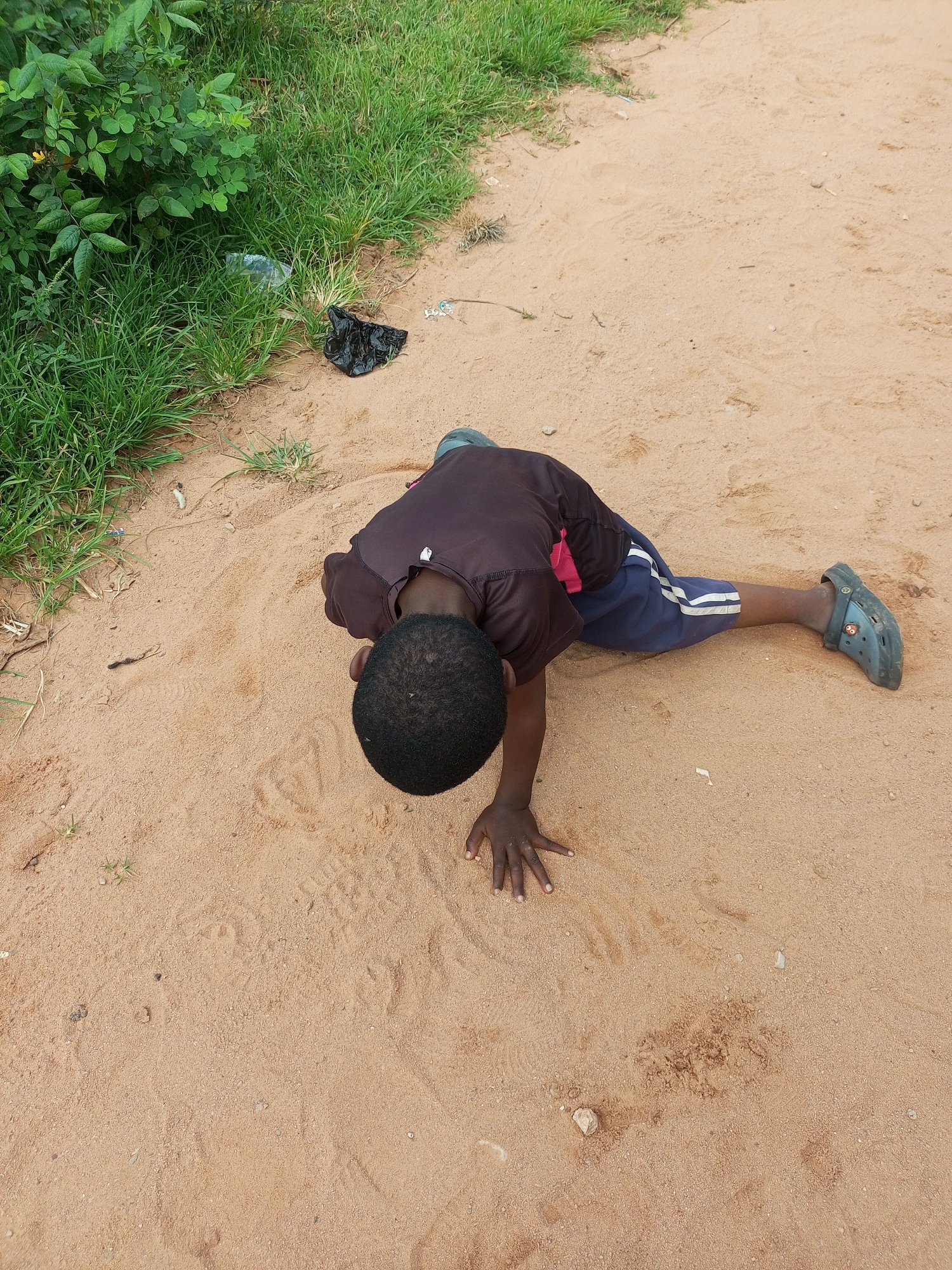 The image shows a young Black boy in the city of Tempure, Nigeria, lying on his stomach on a sandy, dirt path. He is wearing a dark brown t-shirt and blue shorts with white stripes. His left leg is extended behind him, and his right leg is bent at the knee. He is resting on his hands, with his head bowed. There is a patch of green grass and some bushes to the left of the path, where a black plastic bag and a clear plastic bag are visible. The ground is sandy with some small pebbles and vegetation scattered around.
