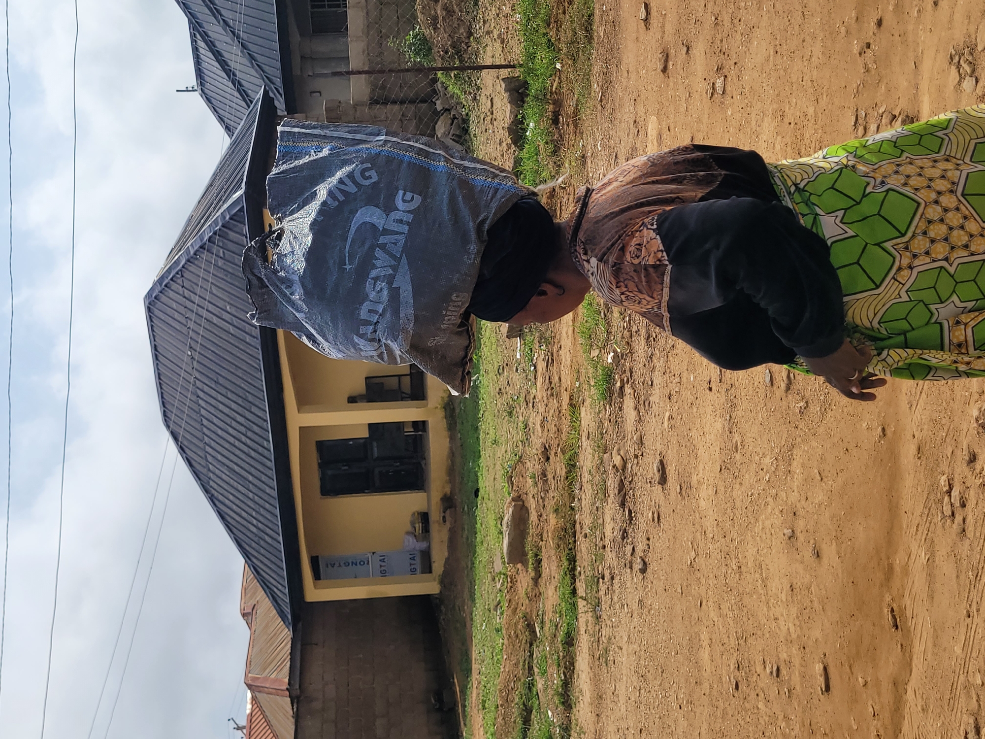 In the city of Jos, Nigeria, a woman walks with a large, dark gray bag of goods balanced on her head. She is wearing a black headwrap, a brown and black patterned top, and a green and white patterned skirt. The background shows a yellow building with a dark roof, a brick wall, and a chain-link fence. The ground is a mixture of dirt and sparse vegetation. The sky is partly cloudy.