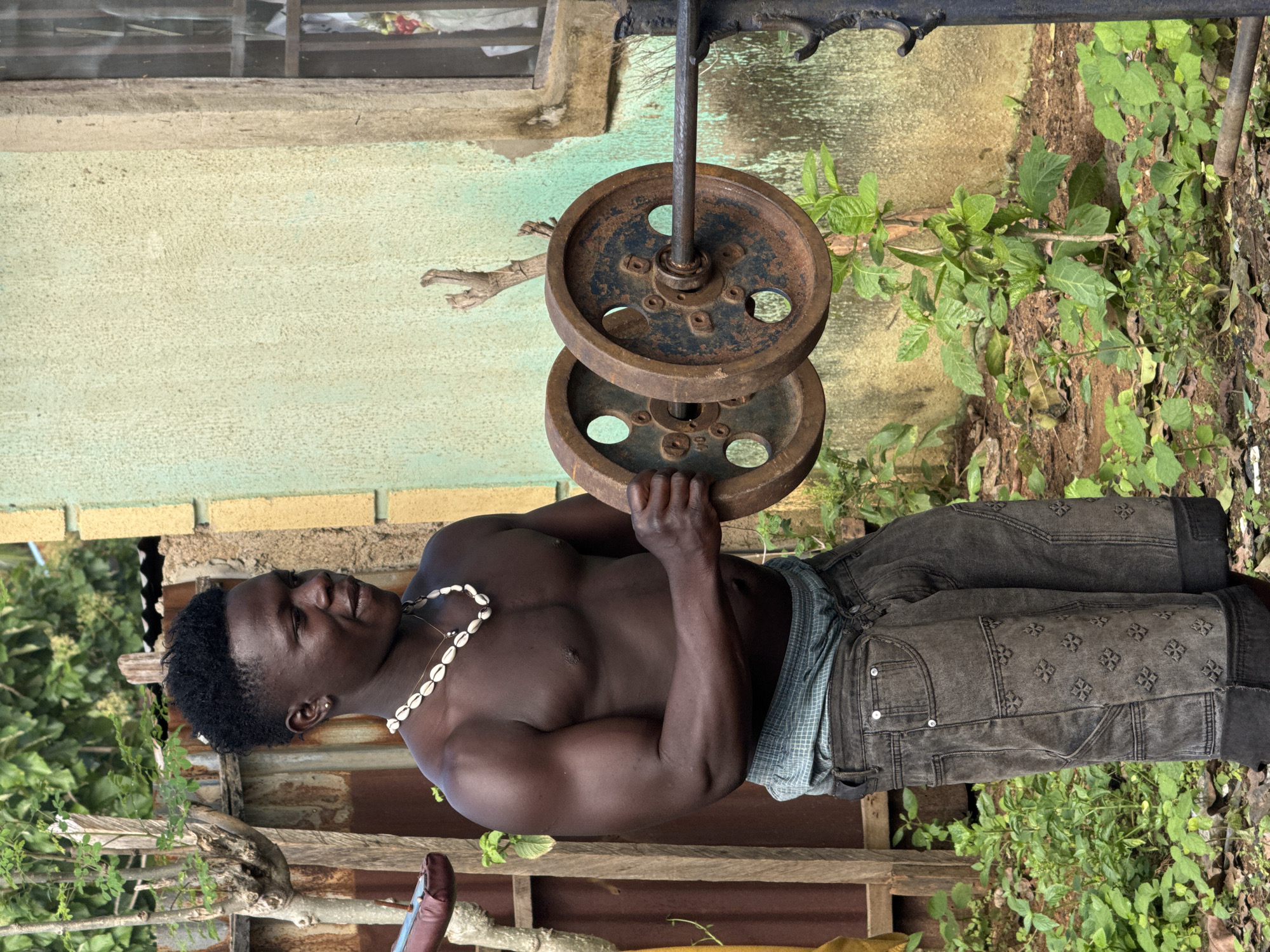 In Akwanga, Nigeria, a shirtless young Black man with dark skin and curly black hair stands outdoors, performing weightlifting exercises. He wears a shell necklace and grey denim shorts. He is holding a barbell with two rusty metal weight plates, one on each side of his hands. The background consists of a textured, pale yellow and green building, a wooden fence, and lush green vegetation.