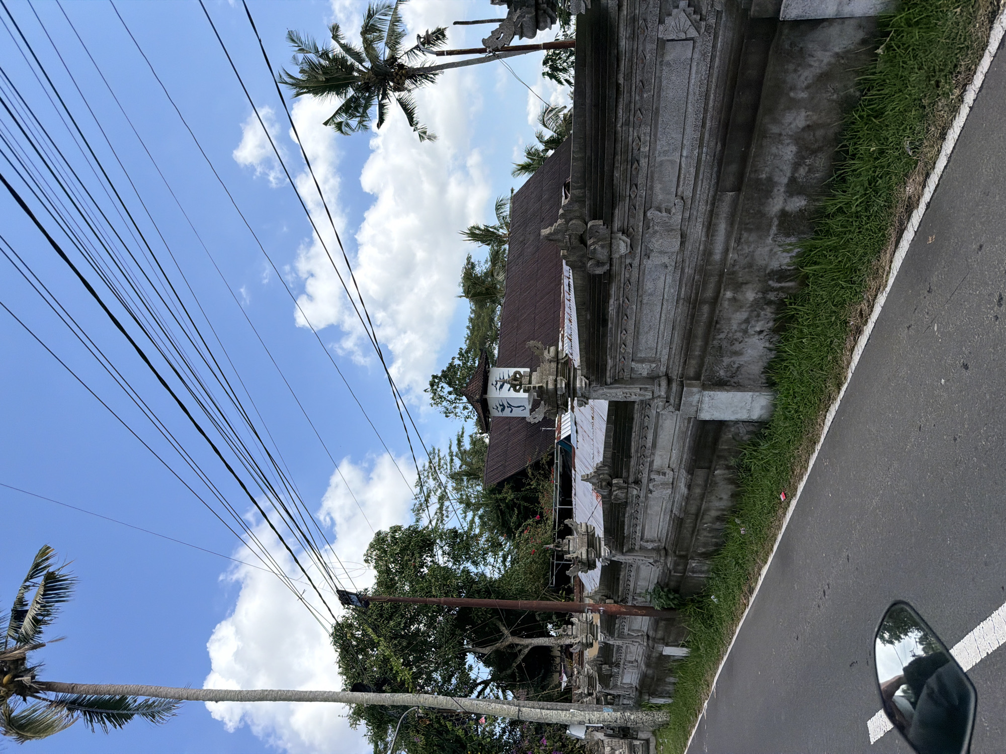 A medium shot captures a street scene in Tampaksiring, Indonesia, under a bright blue sky dotted with white clouds. A tall palm tree stands on the left, its fronds swaying gently. Utility wires crisscross the sky, supported by poles. A grey, ornate stone wall runs along the right side of the road, adorned with intricate carvings. Beyond the wall, the reddish-brown tiled roof of a building is visible, with trees and vegetation surrounding it. The road surface is asphalt, with a white line marking the edge. A motorcycle's rearview mirror is partially visible in the lower left corner, reflecting the sky and trees. The scene appears calm and quiet, suggesting an ordinary day in a tropical locale.