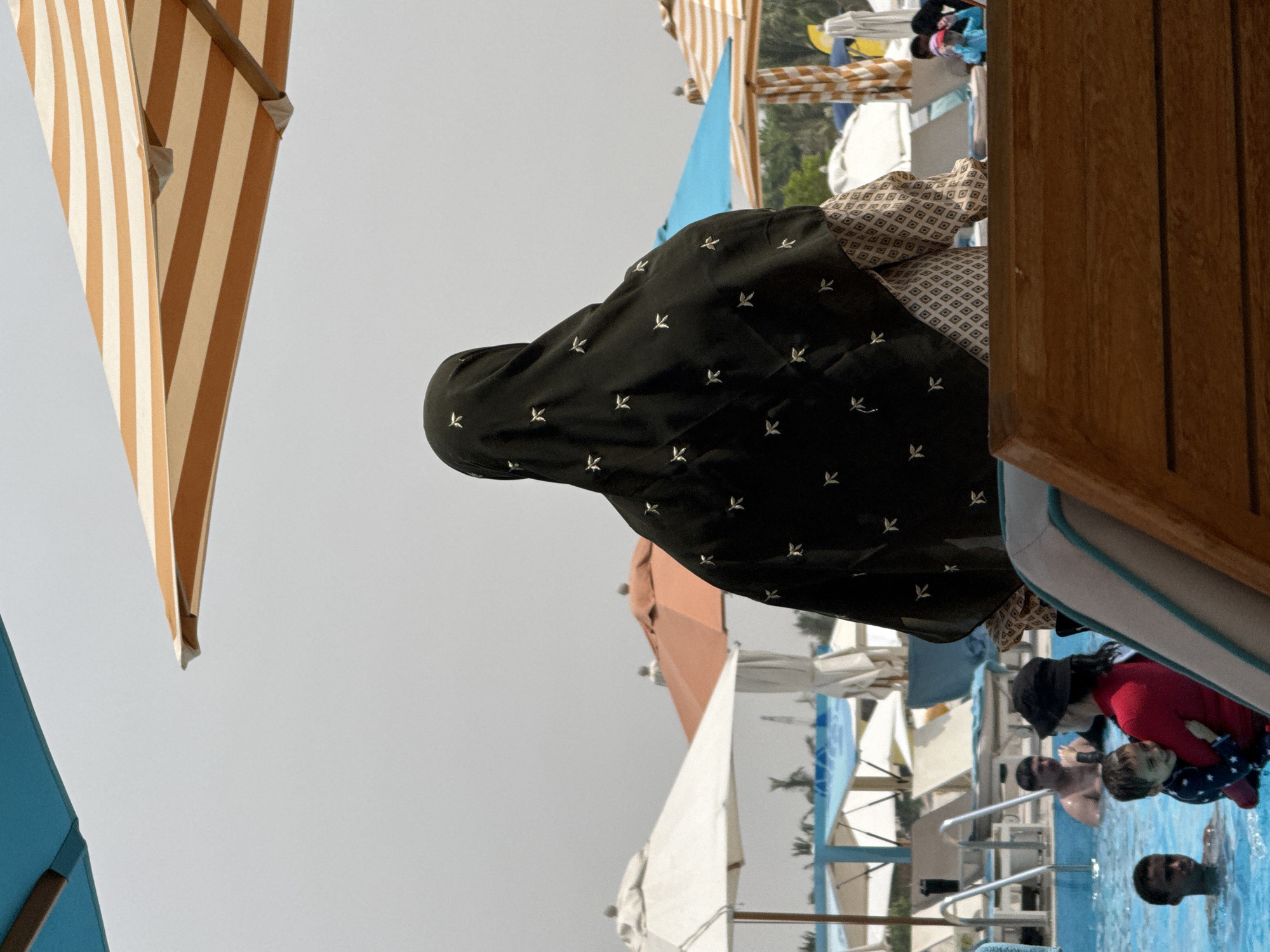 The image depicts a woman wearing a dark patterned headscarf and a patterned dress, sitting on a wooden chair at what appears to be a resort or public pool area. She is viewed from behind. In the background, a swimming pool is visible with several people in it, including children and adults. Umbrellas, some striped and some white, are set up around the pool area, providing shade. The overall environment suggests a sunny day at a recreational facility. The location is identified as Ras Al-Khaimah, United Arab Emirates.