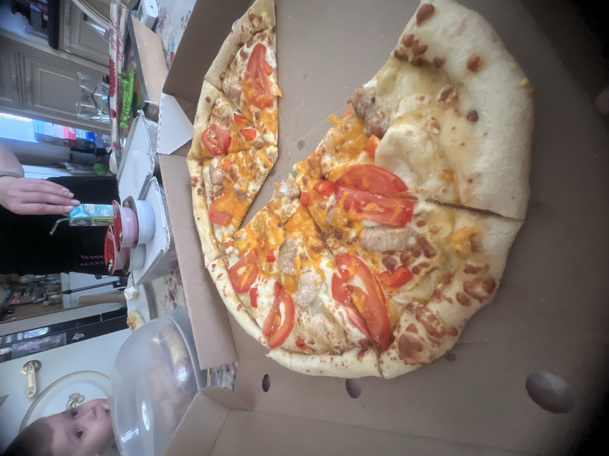 In an indoor, domestic setting, a half-eaten pizza is presented in a cardboard pizza box, positioned on a table with a floral tablecloth. The pizza features a thick crust and is topped with melted cheese, tomato slices, and what appear to be pieces of sausage and bell pepper. To the left of the pizza, a clear plastic dome covers a pastry or bread. A young child, with a curious expression, is visible in the upper left quadrant of the image, looking towards the covered pastry. In the background, a person's hands are visible, holding a juice box. Other items on the table include small bowls, a clear glass, and what seems to be a takeaway food container. The overall lighting suggests an indoor environment, possibly a home, with natural light filtering in from an unseen source.