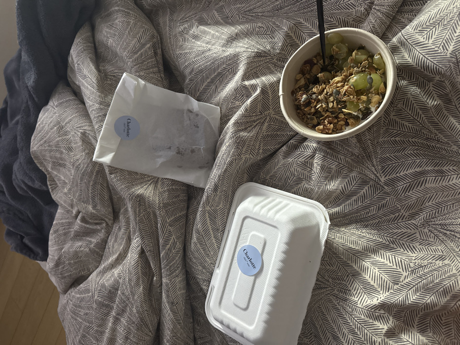 The image displays a meal prepared on a bed, suggesting an indoor bedroom setting. The bed is covered with a rumpled duvet featuring a grey and beige leaf pattern, with a darker blue blanket visible at the top. Sunlight casts distinct shadows across the bedding from an unseen window. Three food items are arranged on the bed. A white, rectangular, lidded takeaway container, possibly made of molded fiber, has a light blue circular sticker on its lid bearing the text "Charlotte. Chleb i Wino." Adjacent to this is a small, crinkled white paper bag, also featuring an identical "Charlotte. Chleb i Wino" sticker, containing a dark, indistinct solid food item. To the lower right, a round, off-white bowl holds a serving of granola, yogurt, and green grapes, with a black spoon partially submerged within. A light brown wooden floor is visible in the upper left background. The "Charlotte. Chleb i Wino" branding indicates the food originates from a "Charlotte. Chleb i Wino" establishment located in Warsaw, Poland.