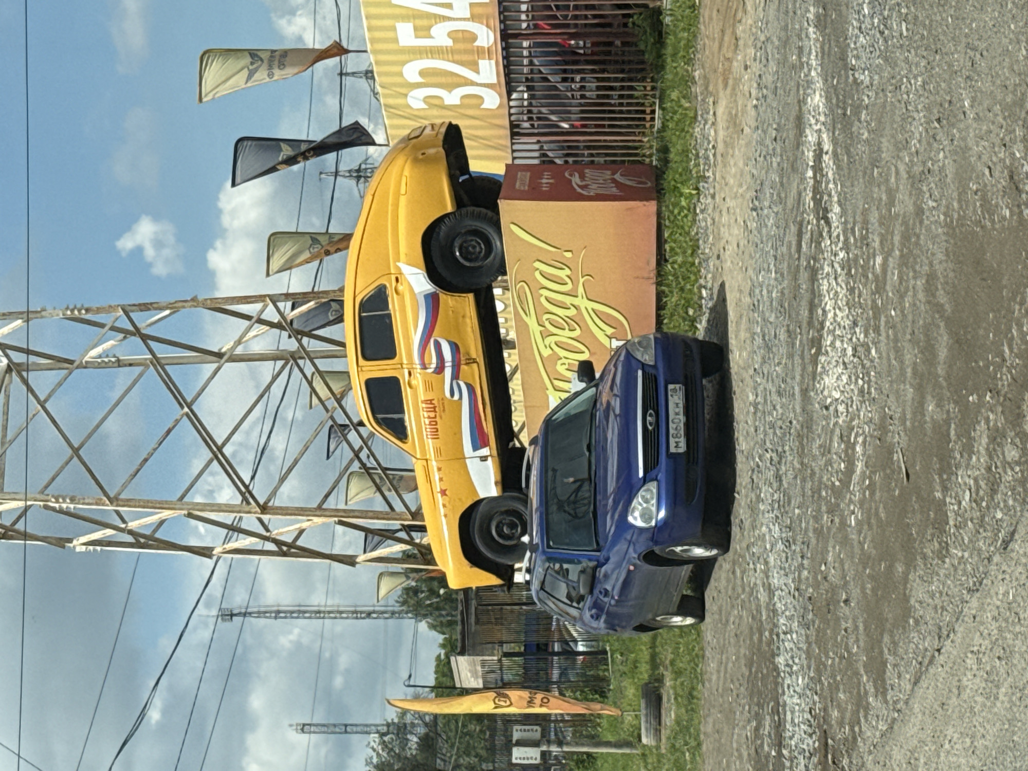 In Izhevsk, Russia, a vintage yellow car, identified as a Pobeda model due to the inscription on its side, is elevated and displayed on top of a blue station wagon. The yellow car is adorned with a red, white, and blue ribbon graphic, symbolizing the Russian flag. The Pobeda has a star emblem on its hood. The blue car, a Lada Granta, is parked on a gravel lot directly beneath the elevated car. Behind the vehicles, a large industrial tower structure stands prominently. Several tall, thin flags, some displaying emblems and text, are also visible in the background, along with a yellow advertising banner featuring the digits "3254". The scene is set against a cloudy sky with patches of blue.