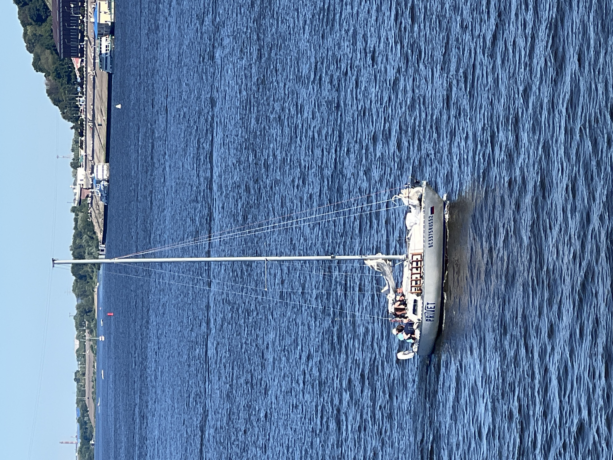 A sailboat with "PRIVET" and "BC 3375 RU52" written on its hull is sailing on the Volga River in Nizhny Novgorod, Russia. Several people are visible on the deck of the sailboat. In the background, the cityscape of Nizhny Novgorod is visible, including buildings and a hill. The sky is clear and blue.