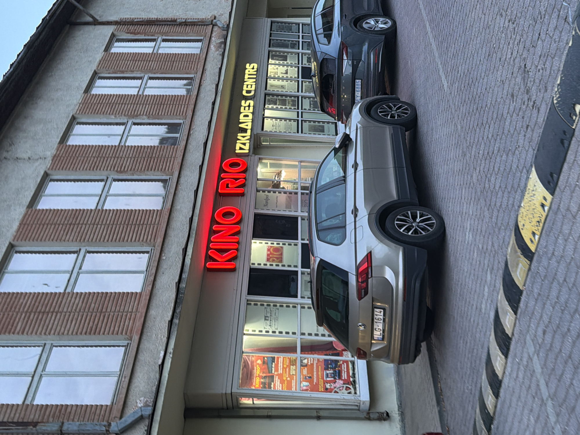 The image shows the facade of a building with the neon sign "KINO RIO" in red and "IZKLAIDES CENTRS" in yellow. The building appears to be a cinema and entertainment center. In front of the building, two cars are parked on a cobblestone surface. The car in the foreground is a light brown Volkswagen Tiguan with the license plate "LG-1614". The car in the background is a dark grey Skoda station wagon with the license plate "EY-6885". A speed bump is visible in the lower part of the image. The building has large windows displaying movie posters and what appear to be framed images. The architecture suggests a slightly older building, possibly in a European city. Based on the signage and the license plate format, the location is likely Ventspils, Latvia, a city known for its cinema named Kino Rio.