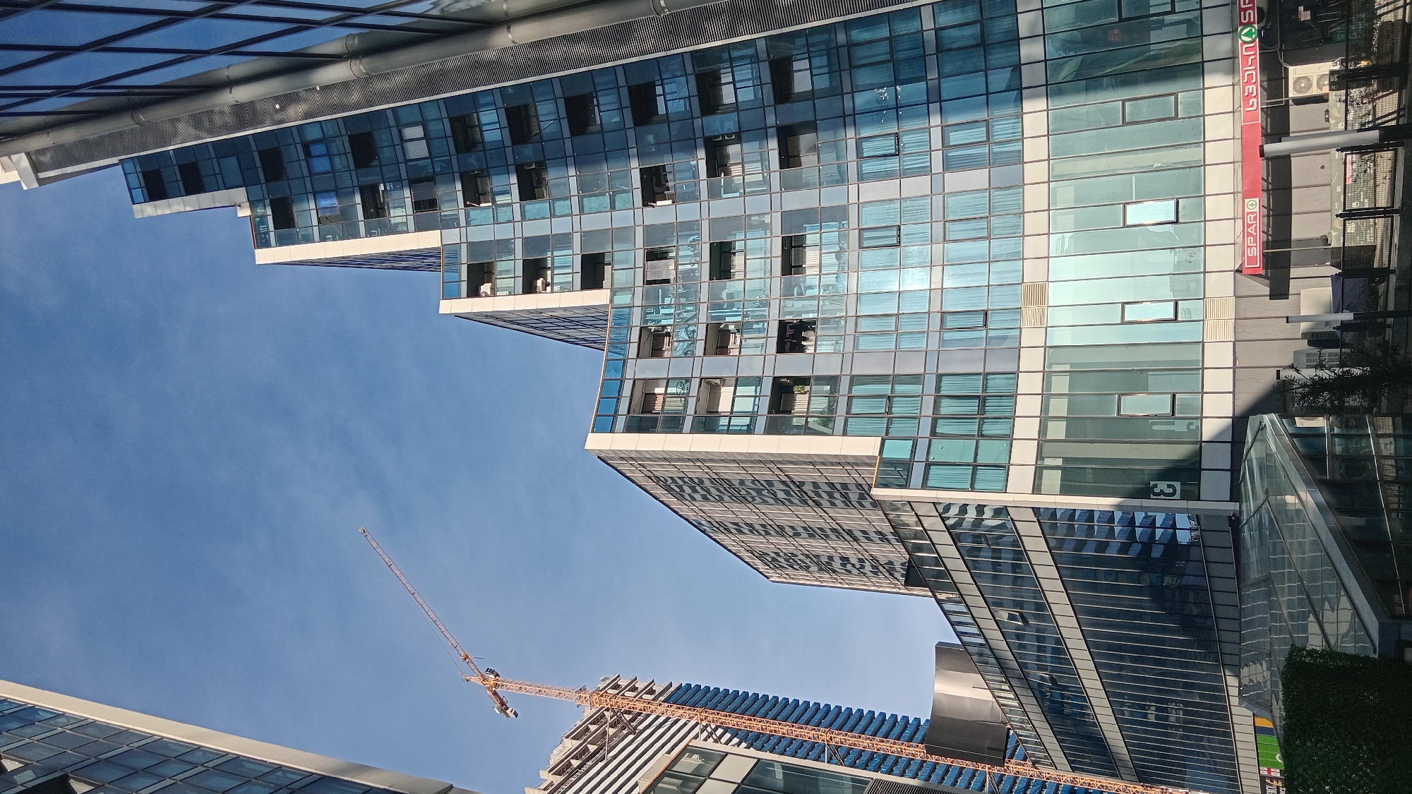 The image is a low-angle shot looking up at modern buildings with glass facades under a clear blue sky. A construction crane is visible in the lower portion of the frame. The reflection of the sky and other buildings is captured in the glass windows. A commercial sign with the word "SPAR" is visible in the upper right side of the frame. The image depicts an urban environment in Batumi, Georgia.