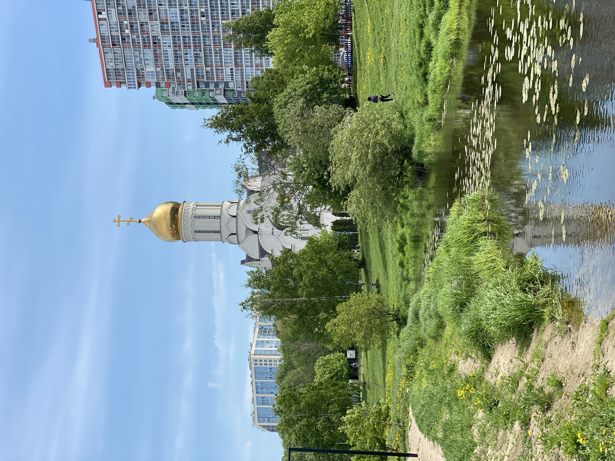 The image shows a cityscape with a white church and golden dome juxtaposed against modern high-rise buildings. The church is positioned towards the left, its reflection visible in a body of water to the right. The water contains lily pads. A grassy bank separates the water from a walking path. People are seen in the background, near the high-rise buildings. The sky is blue with light clouds. The location is in the city of Unknown, Unknown.