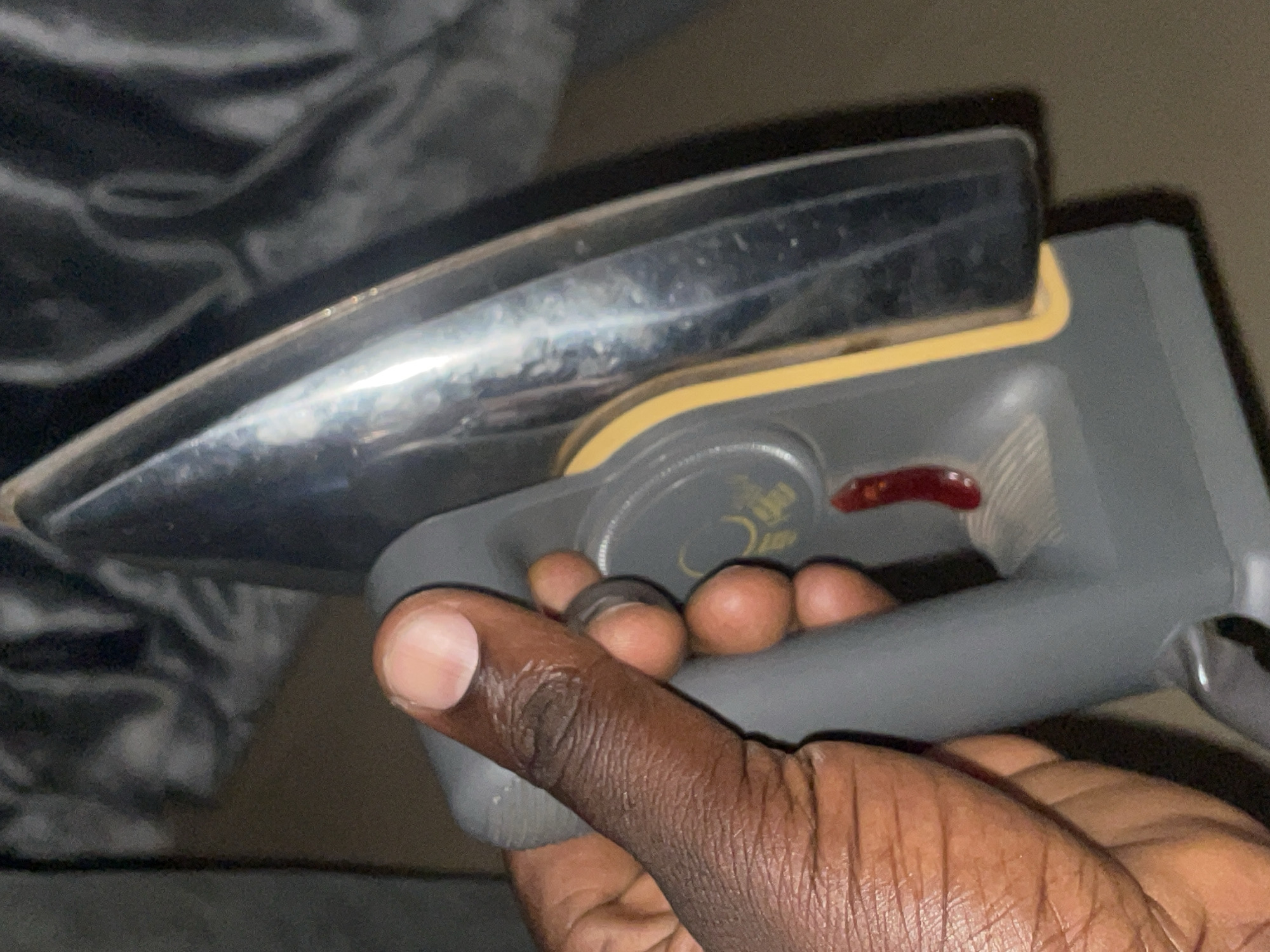 An individual's hand, showing skin texture and fingernails, is gripping the grey handle of an iron. The iron's metallic soleplate is visible at an angle, with a yellow accent bordering its edge. A red indicator light is present on the iron's body, near a circular dial with faint markings. The background is blurred and dark, suggesting an indoor setting. The context of Jos, Nigeria, is not discernible from the visual information provided in the image.