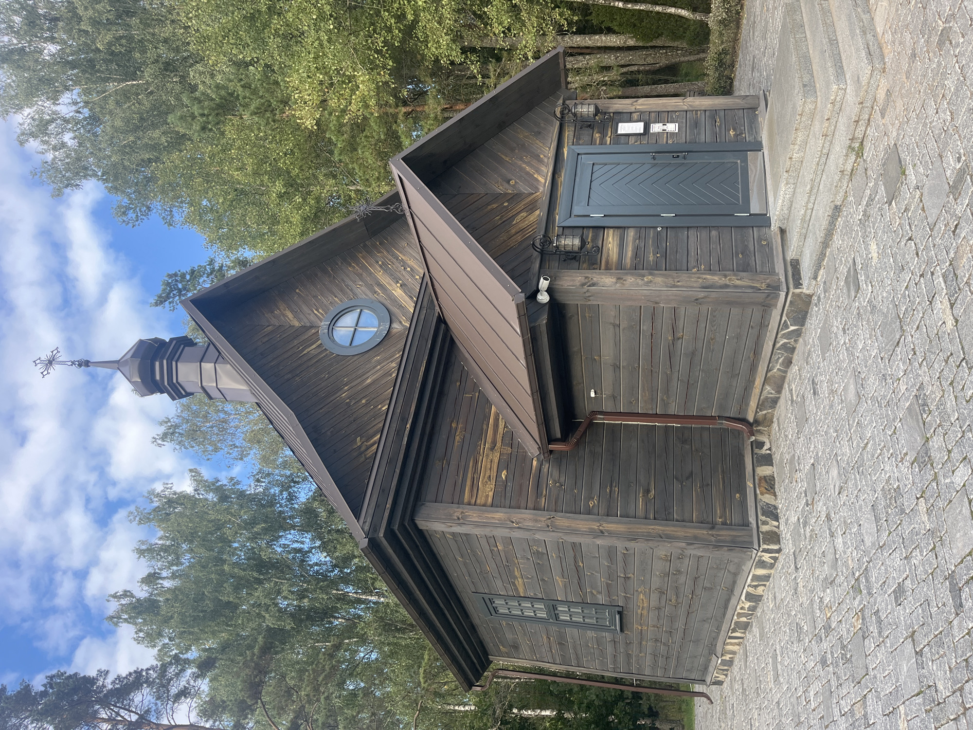 The image shows a small, dark wooden church with a bronze-colored roof. The church has a simple, rectangular structure with a gabled roof that rises to a small steeple topped with a cross. There is a circular window above the entrance and a long, narrow window on the side. The entrance is recessed and has a single door with a chevron pattern. Two outdoor lanterns flank the door, and a security camera is mounted above them. The church is surrounded by trees, with a paved walkway leading to the entrance. The paving stones are a mix of gray and black, creating a mosaic pattern. The overall scene is quiet and serene, suggesting a place of worship in a natural setting.