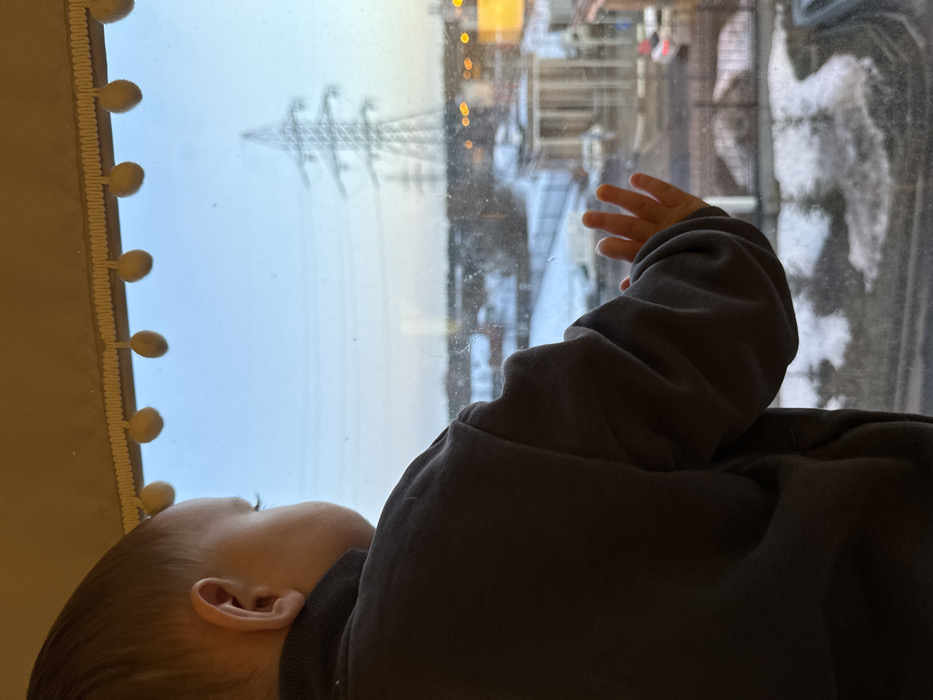A young child, observed from behind and the right side, looks out a window in Akulovo (Gp Kubinka), Russia. The child has light brown hair and wears a dark long-sleeved garment. Their right hand is pressed flat against the windowpane. Above, a light-colored valance with spherical pom-poms decorates the window frame. The exterior scene, viewed through the window, depicts an urban or semi-urban environment with patches of snow on the ground. A street, buildings, and vehicles are visible. Multiple large metallic electricity pylons stand prominently in the distance against a pale blue sky, suggesting either early morning or late afternoon light. Distant artificial lights are visible.