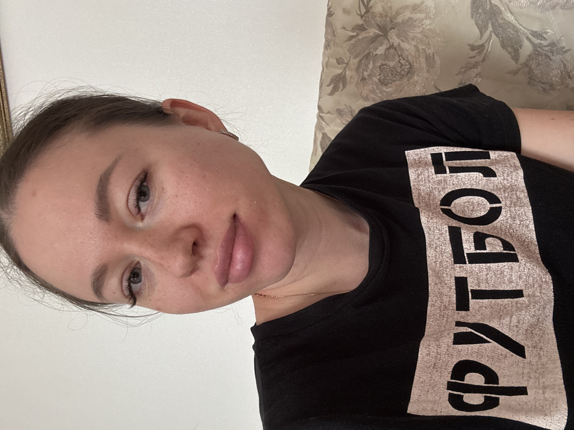 A woman is shown in a close-up, angled selfie. She has fair skin, light brown hair pulled back, and appears to be wearing minimal makeup. Her t-shirt is black with a light-colored inscription in Russian that translates to "football" or "soccer". The background consists of a plain light-colored wall on the left and a textured floral pattern on what seems to be a sofa or upholstered headboard on the right. There is no clear evidence to place her in Ферма, Russia.