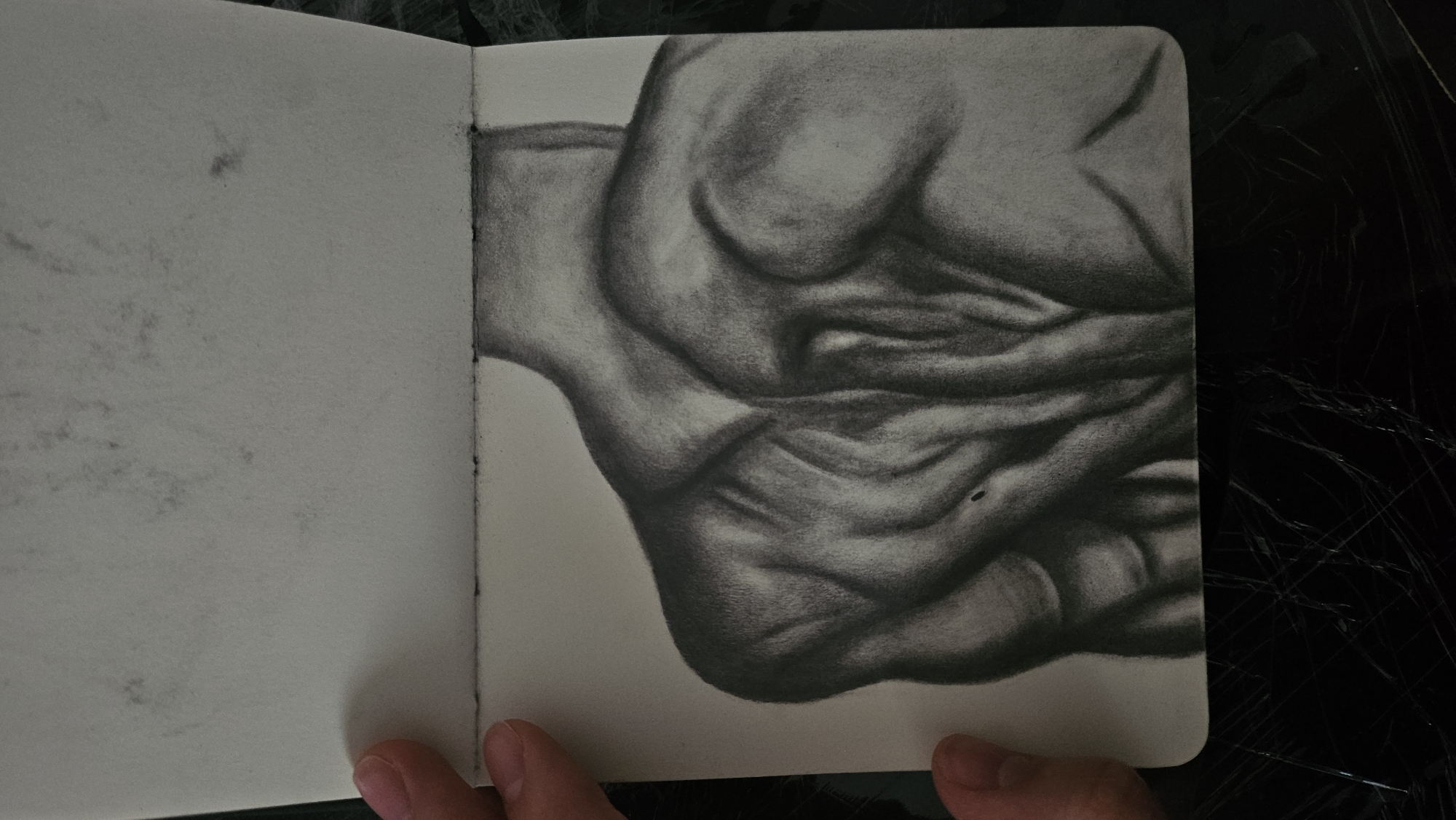 A charcoal drawing on paper depicts the upper back and shoulders of a muscular individual. The drawing is of a dark-skinned male. The muscles are highly defined with deep shadows and highlights, showcasing anatomical detail. The drawing is presented in an open sketchbook. The top page of the sketchbook shows smudges and scuff marks. The lower page, containing the drawing, has a smooth, light gray surface. The edges of the paper are slightly uneven and show some darker smudges, possibly from the charcoal medium. A person's fingers are visible on the left side, holding the sketchbook open. The background appears to be a dark, textured surface. The setting is indoors, as indicated by the consistent lighting and the sketchbook format. No specific location within a city is identifiable.