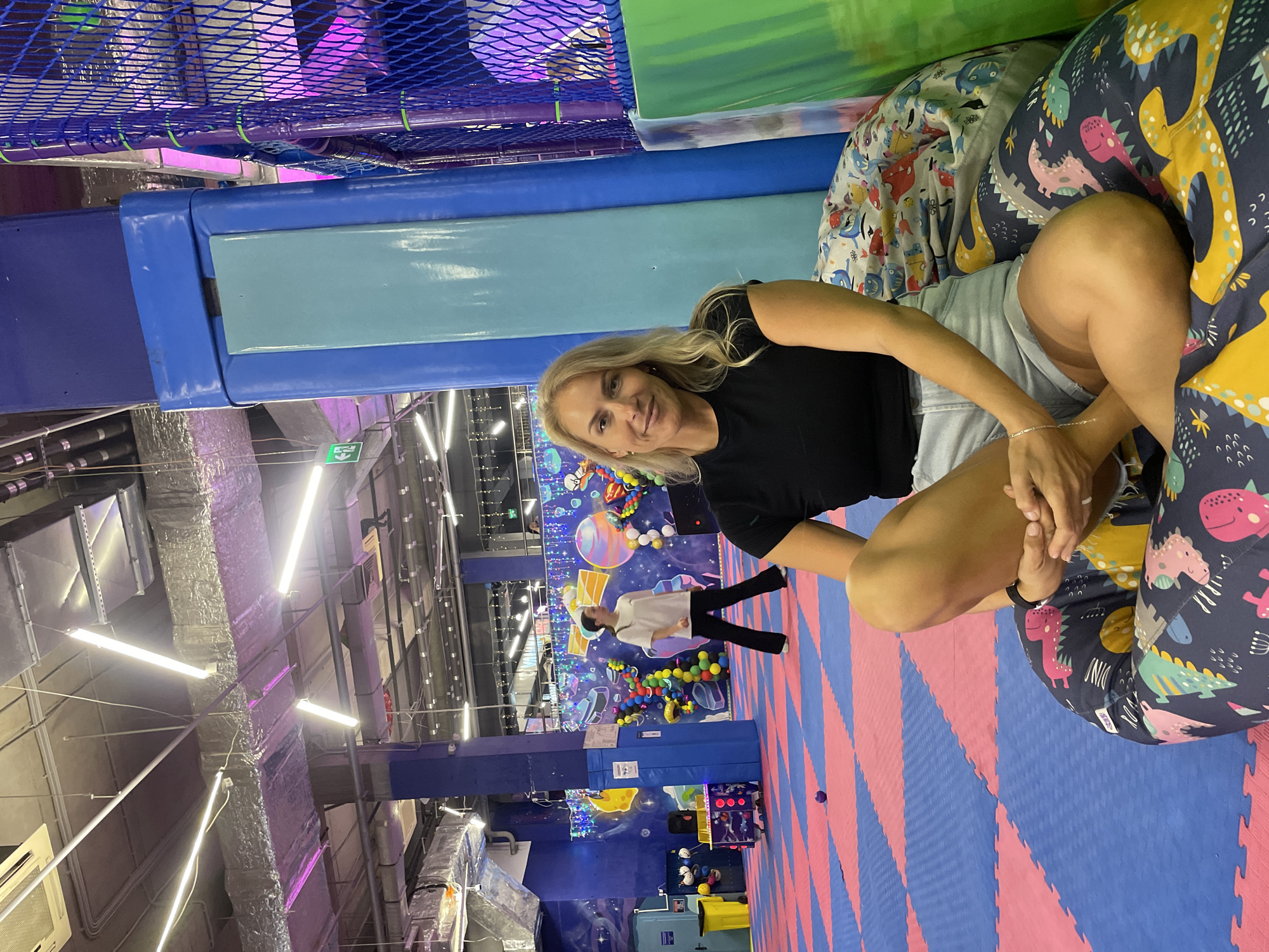 The image shows a blonde woman sitting on a beanbag chair in what appears to be an indoor playground or entertainment center. She is wearing a black t-shirt and light-colored shorts. The beanbag has a dinosaur pattern on it. The background includes playground equipment, colorful flooring, and decorations with a space or planetary theme. Fluorescent lights are visible overhead. A person is walking in the background near the space-themed decorations. The exact location in Sochi, Russia, is not discernible from the image.