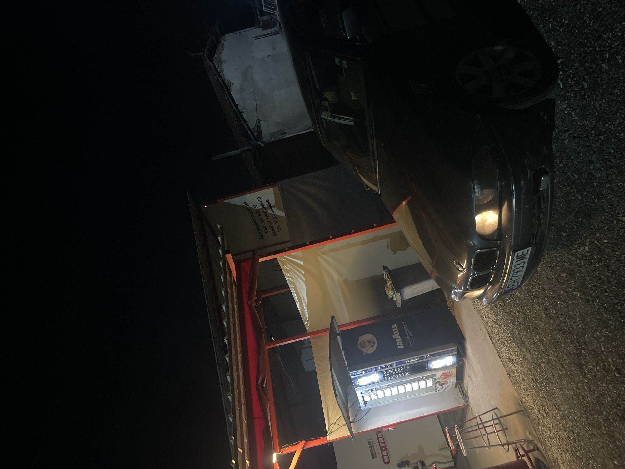 In the city of Dolni Dabnik, Bulgaria, at night, a dark-colored BMW sedan is parked on a gravel lot. The car's headlights are illuminated, and its license plate reads "EH 7933 ME". Adjacent to the car is a covered structure with a red frame and a corrugated metal roof. Inside this structure, a tall vending machine, branded with "LAVAZZA ITALY'S FAVOURITE COFFEE", is dispensing drinks. To the left of the vending machine, a red and white stool is visible. Behind the vending machine, a trash receptacle with some items on top is situated. A partially visible sign with Bulgarian text is mounted on the wall of the structure. Further in the background, a dilapidated building with a damaged roof is seen. The overall scene suggests a commercial area or a rest stop operating after dark.