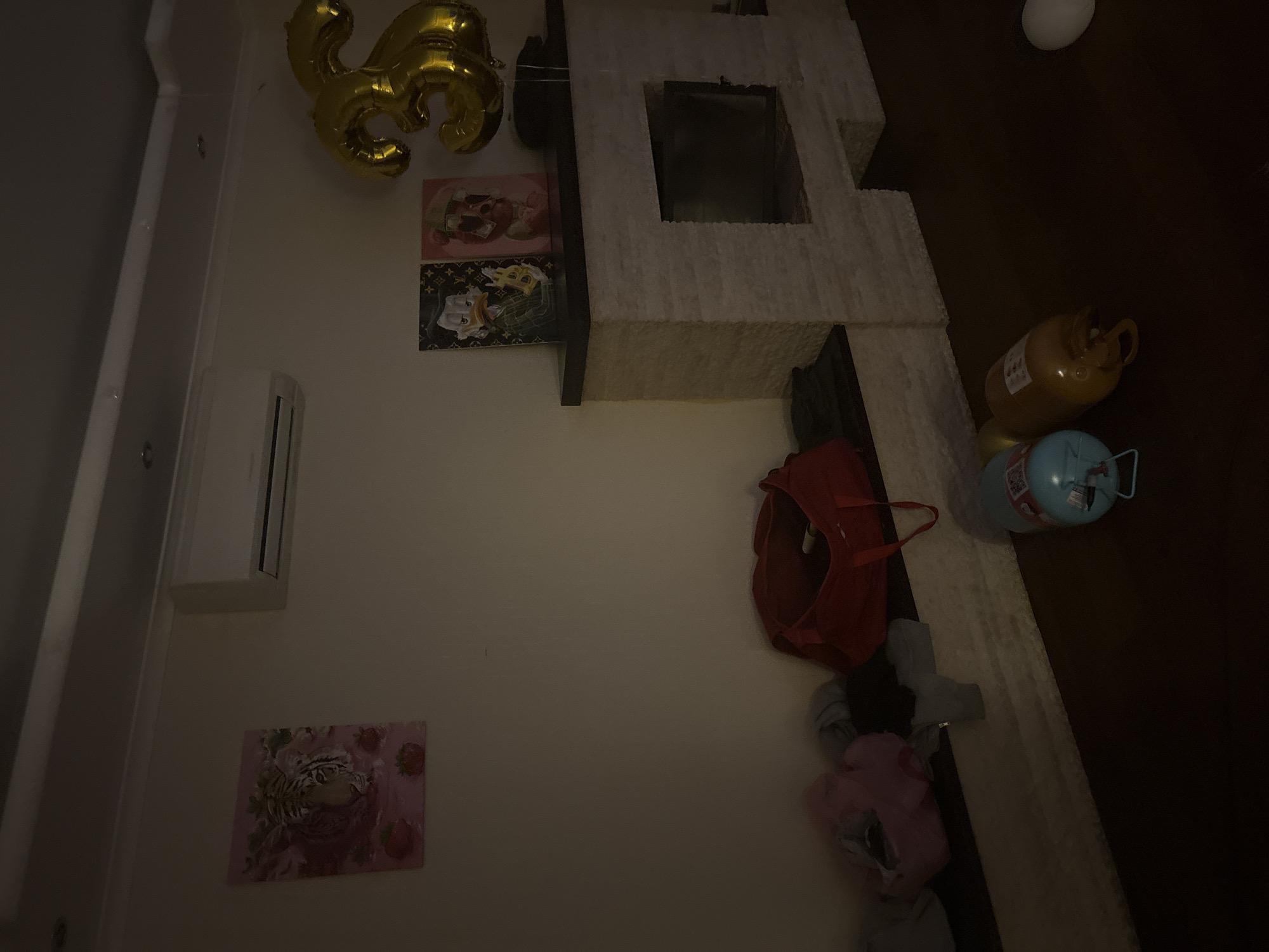 The image depicts an indoor scene with a focus on a wall decorated with artwork and balloons. On the left wall, a painting of a tiger's head surrounded by strawberries is displayed, with an air conditioning unit mounted above it. To the right of the tiger painting, two framed pictures featuring cartoon characters, one resembling Donald Duck and another a bear, are placed on a mantelpiece. A large, gold "3" shaped balloon floats above the mantelpiece. The mantelpiece itself is constructed of white stone blocks. Below the mantelpiece, on a low shelf, are several bags and clothing items, including a red duffel bag. On the floor in front of the mantelpiece are two propane tanks, one blue and one yellow, and a white balloon. The room appears to be dimly lit. The scene suggests a domestic setting, possibly decorated for a celebration.