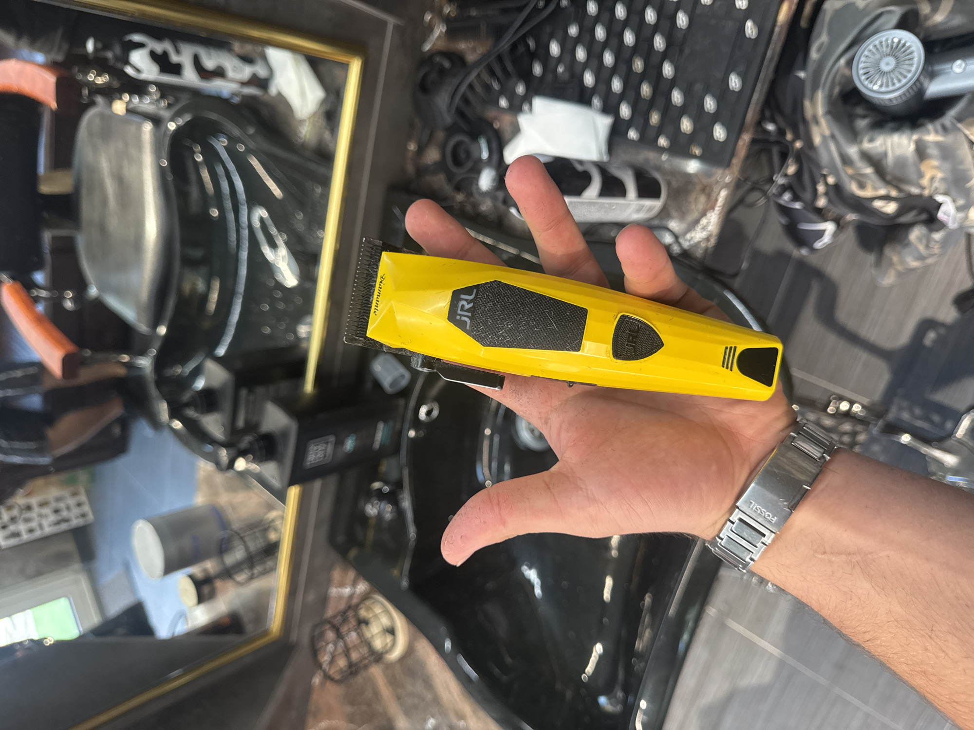 This is a close-up, first-person perspective image set in what appears to be a barbershop or hair salon in Varna, Bulgaria. The central subject is a person's hand, likely male, holding a bright yellow hair clipper. The clipper has "JRL" and "Trumanto" printed on its side, along with a black textured grip.
The hand is adorned with a silver-colored metal watch band, clearly displaying the brand name "FOSSIL" engraved on it. The background provides context for the setting. To the left, a mirror reflects various elements of the salon, including a black barber chair, a sink or basin, and a glimpse of what appears to be an outdoor scene with green foliage through a window. Directly behind the hand and to the right, a black textured mat or tray with numerous circular indentations is visible, along with a partially seen gray hairdryer and some white tissues. Below the hand, another black sink or basin is present, along with a power adapter or control box and a wire basket on the floor.
No specific activity is taking place other than the hand holding the clipper, possibly for display or a product shot. The lighting suggests it is daytime, with both natural light from a window and artificial indoor lighting illuminating the scene. No explicit emotions are conveyed, as only the hand is visible.